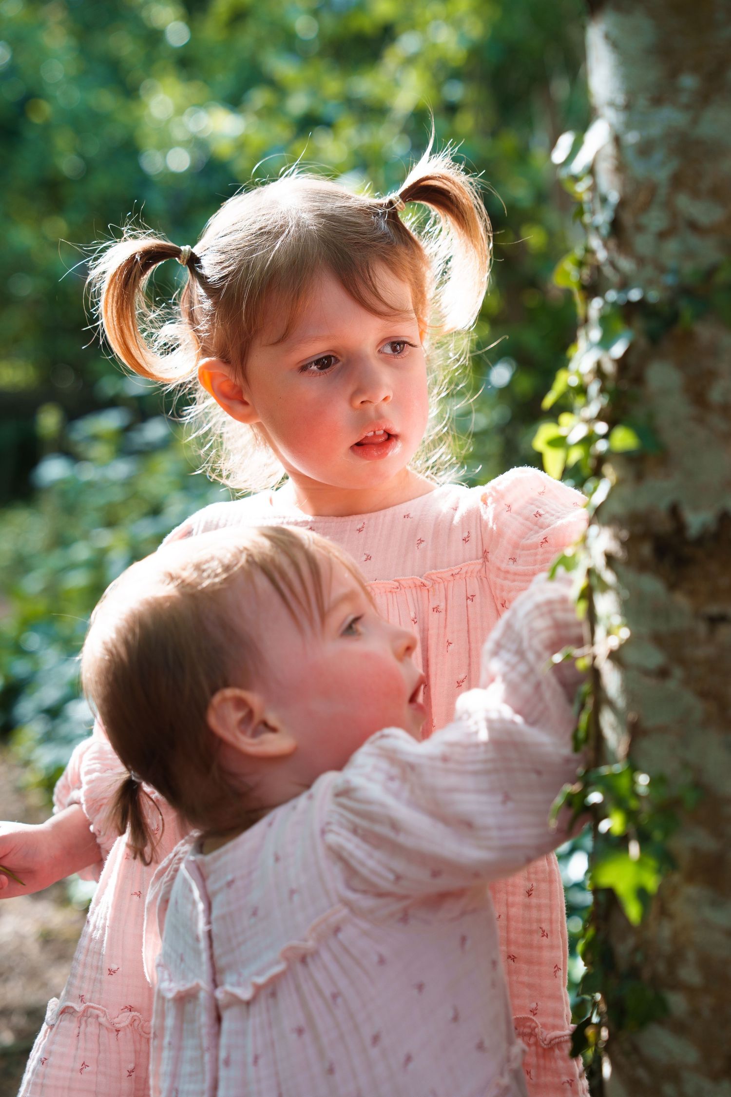 Two young girls in pink dresses playing near a tree in a sunny garden, one with pigtails looking curiously ahead.