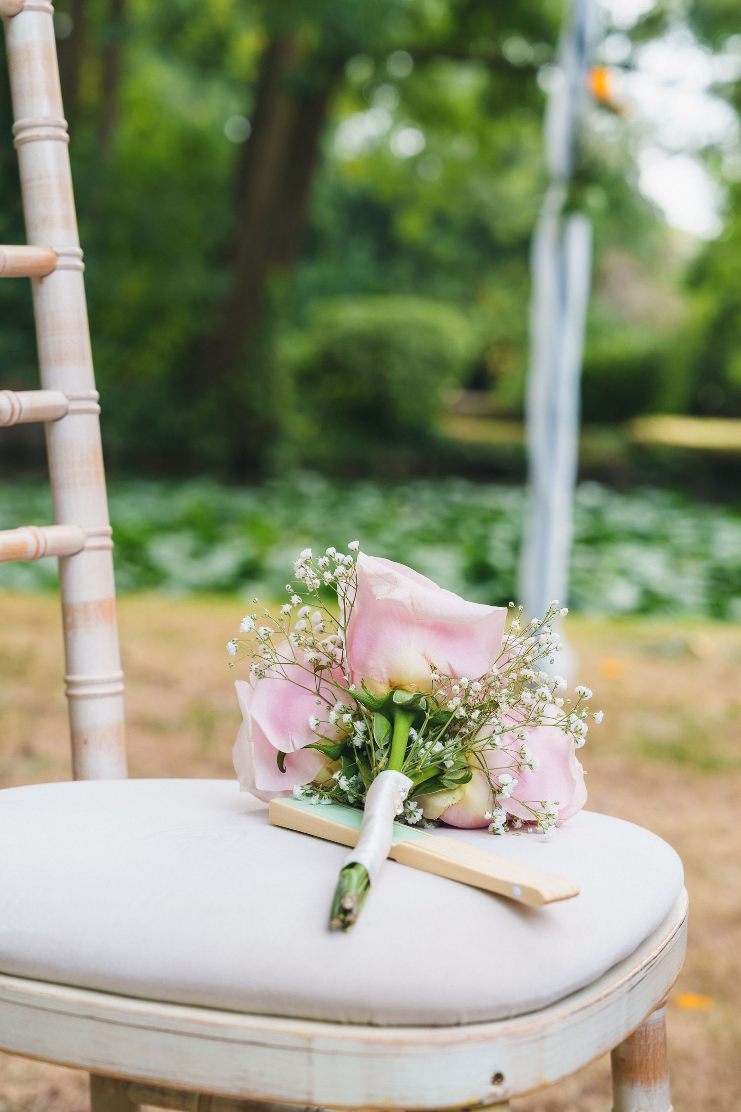 Pink rose and baby's breath bridal bouquet resting on a white chair at an outdoor garden wedding ceremony.
