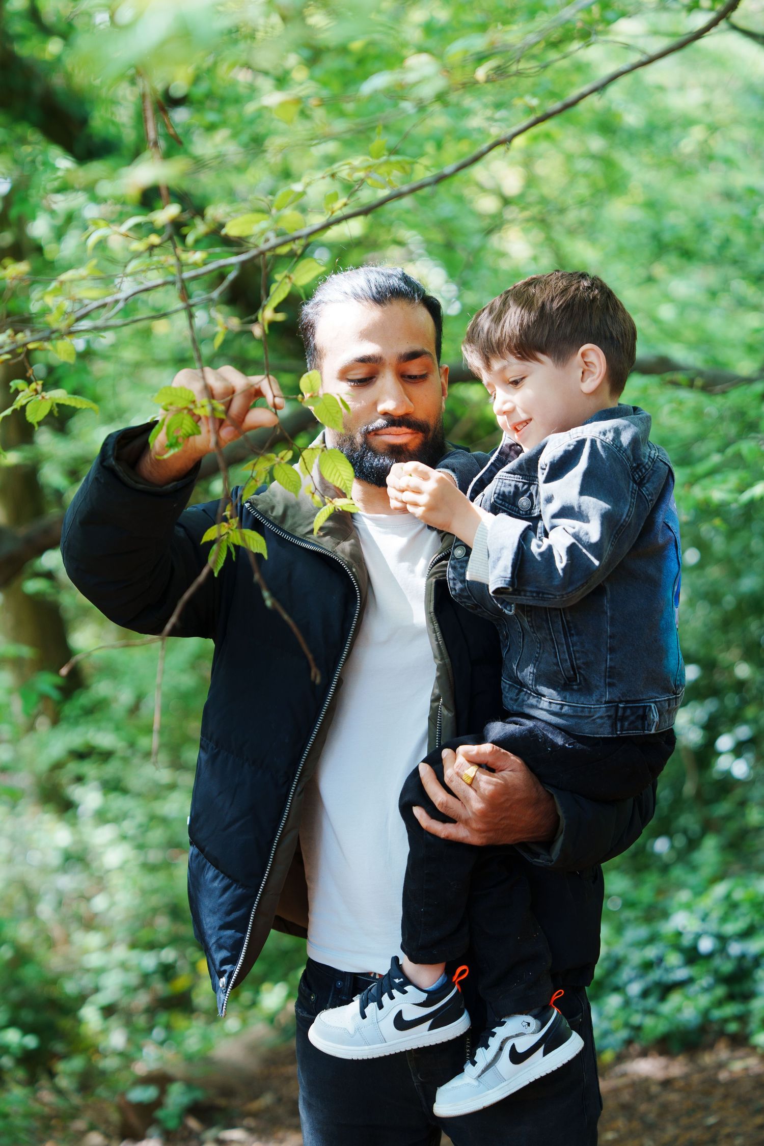 A bearded man holds a young boy while exploring green forest trees, both wearing black jackets and Nike sneakers outdoors.