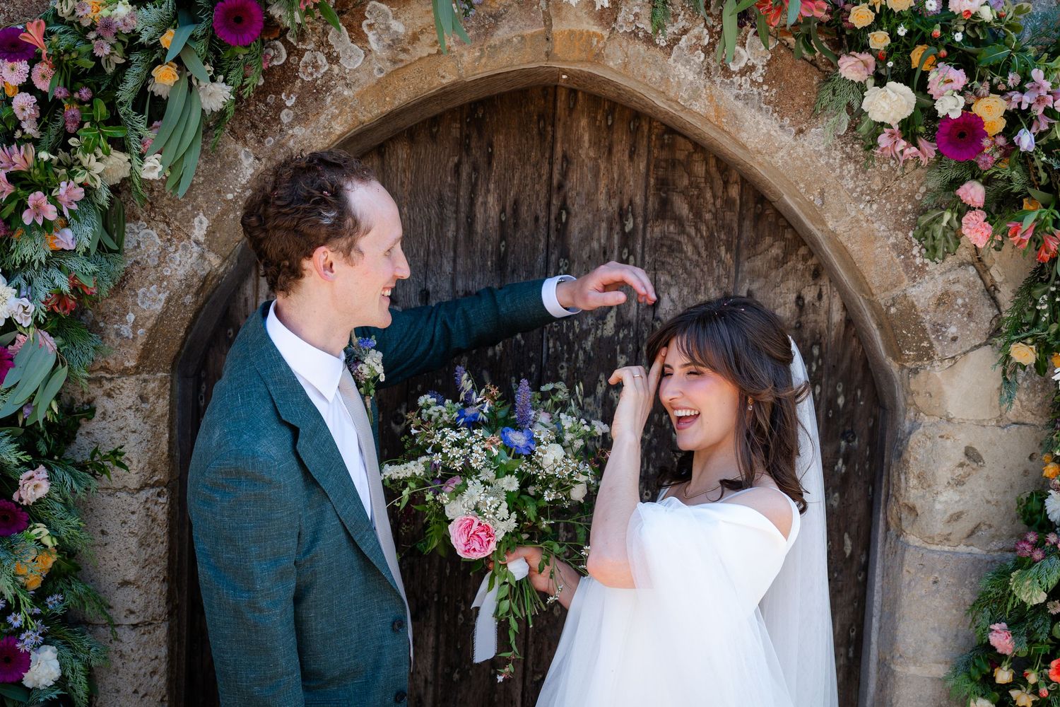 Groom in teal suit playfully twirls bride's hair under floral arch at rustic church wedding.