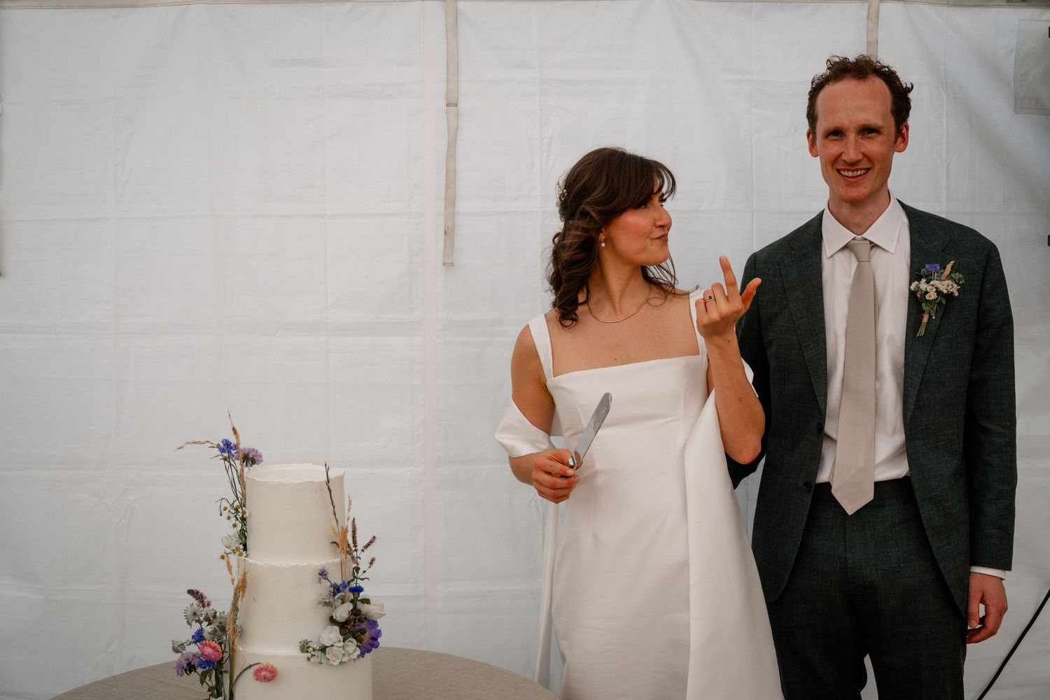 Bride and groom smiling while cutting their white wedding cake decorated with wildflowers in a marquee tent.