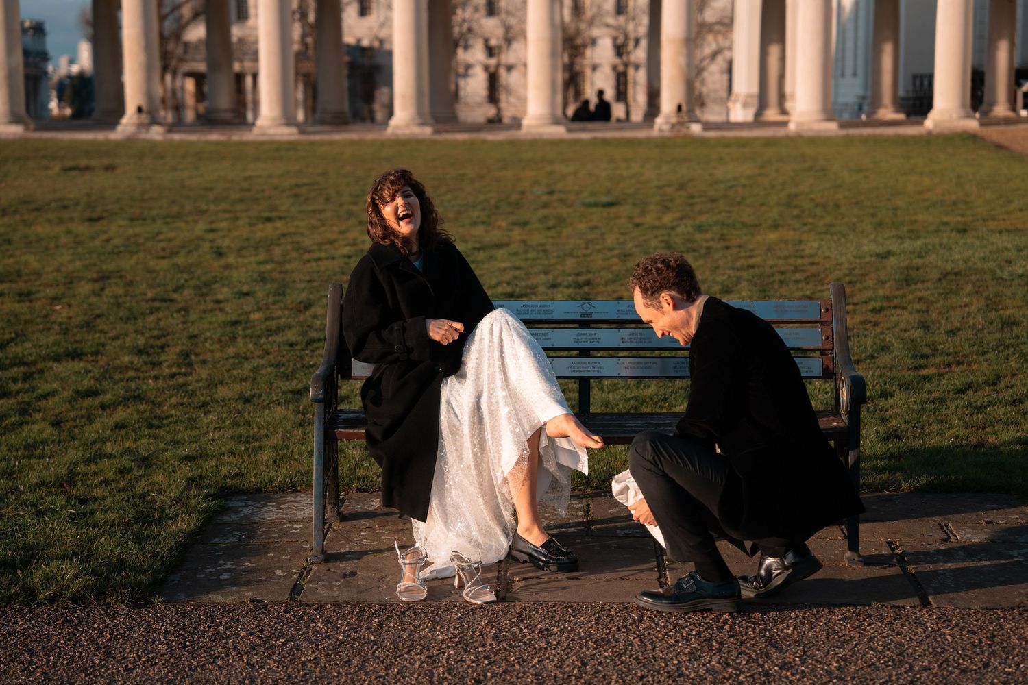 A couple sits on a park bench with classical columns in the background during golden hour.