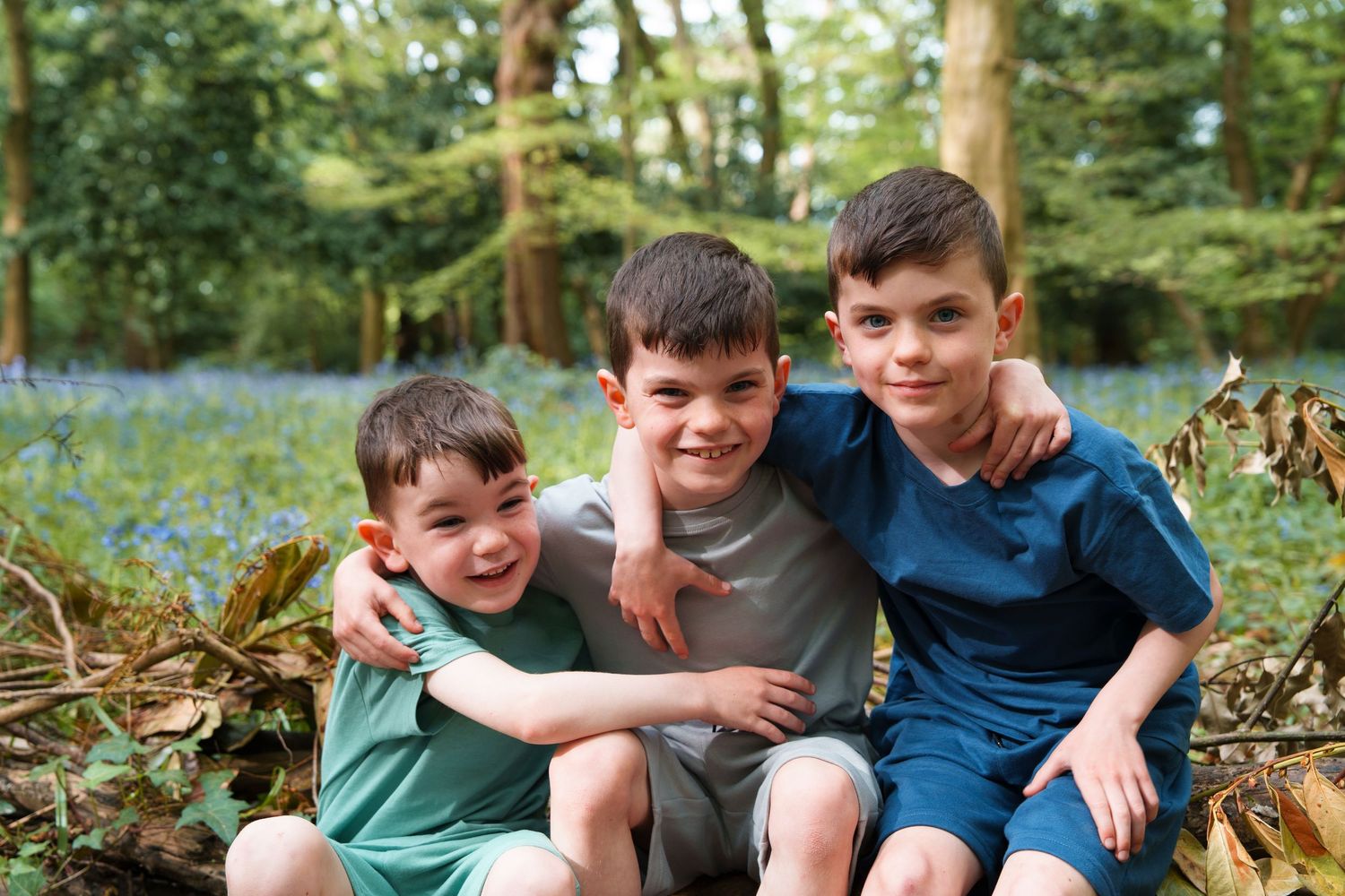 Three young boys smiling and hugging in a bluebell woodland, sitting together outdoors among trees and autumn leaves.