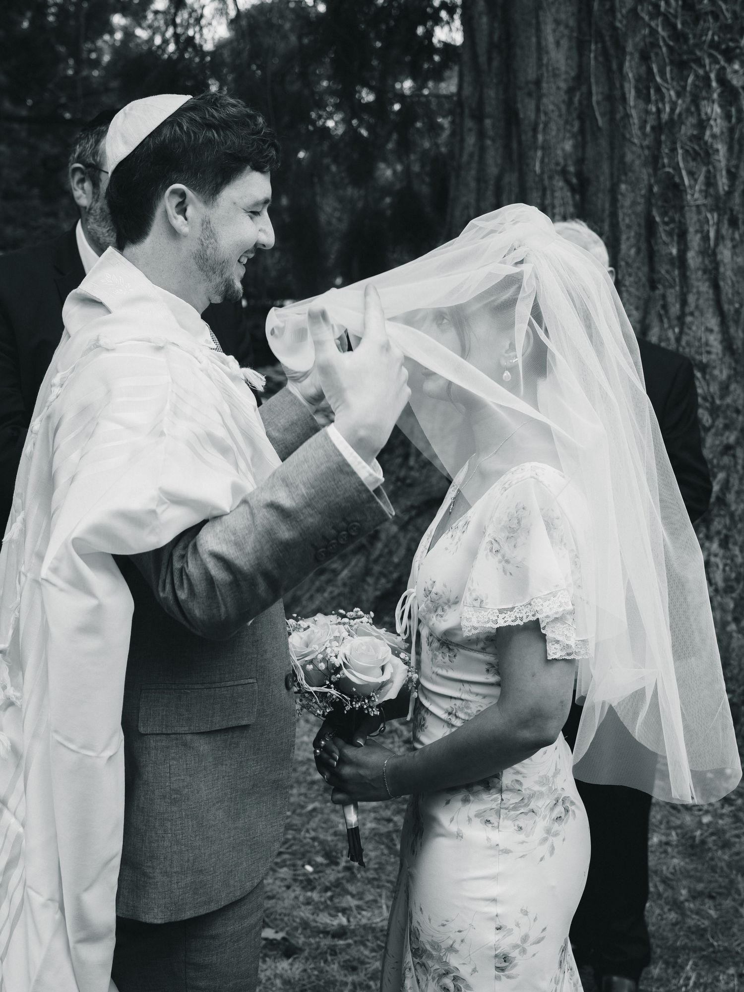 Groom lifting bride's veil during Jewish wedding ceremony outdoors, black and white photo.