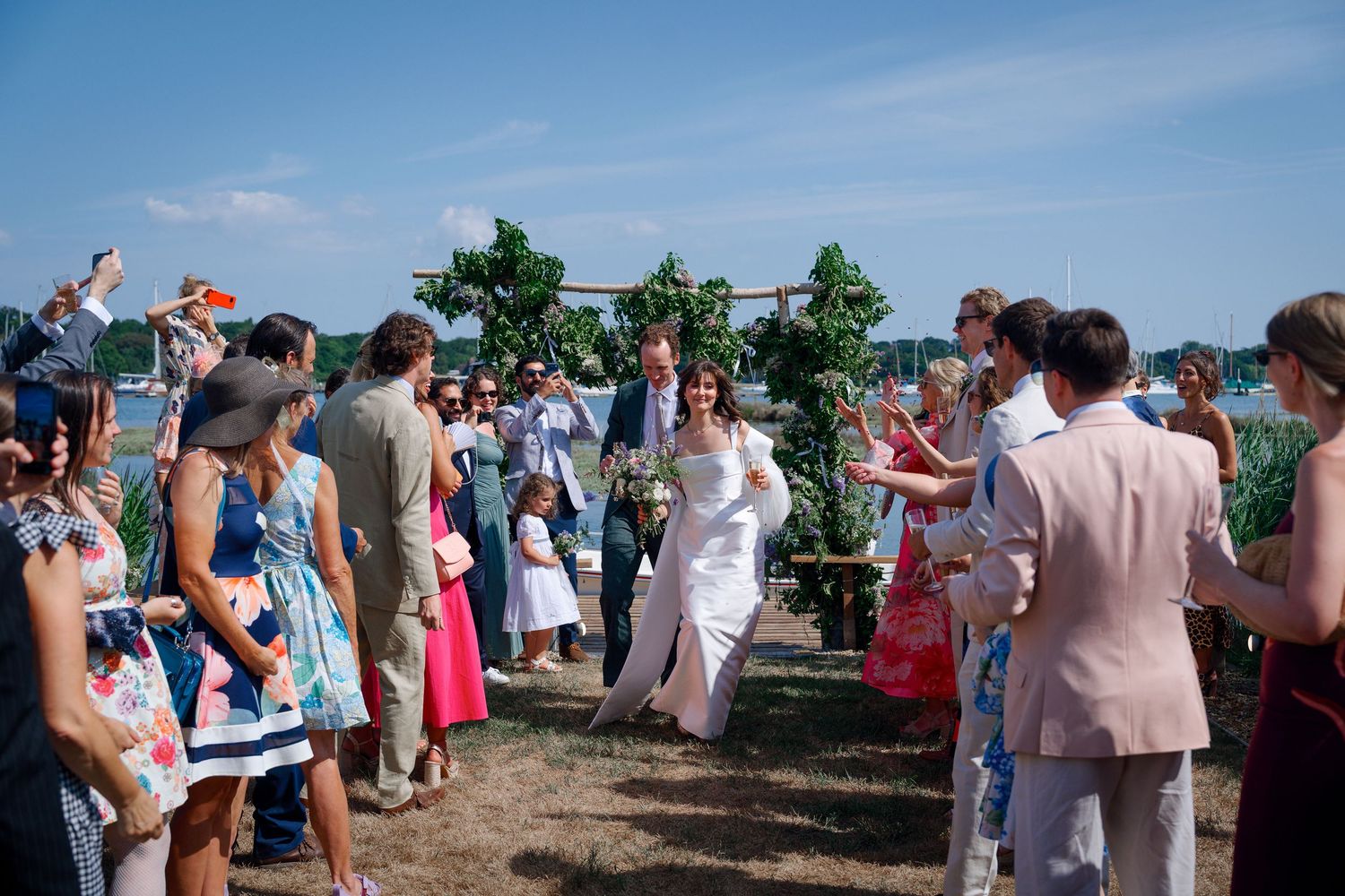 Bride and groom walk down outdoor aisle as guests celebrate with confetti at waterfront wedding ceremony.