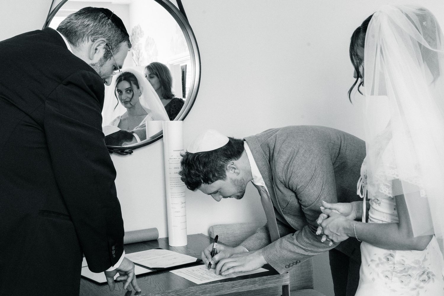 Groom signing Jewish wedding ketubah contract at ceremony table with bride and officiant in black and white photo.