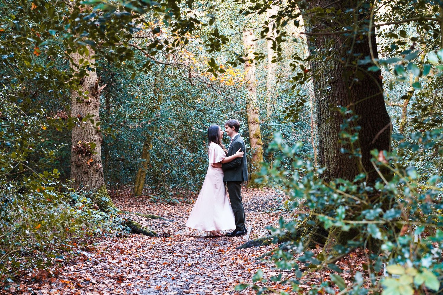 A couple embraces on a leaf-covered forest path surrounded by lush greenery.