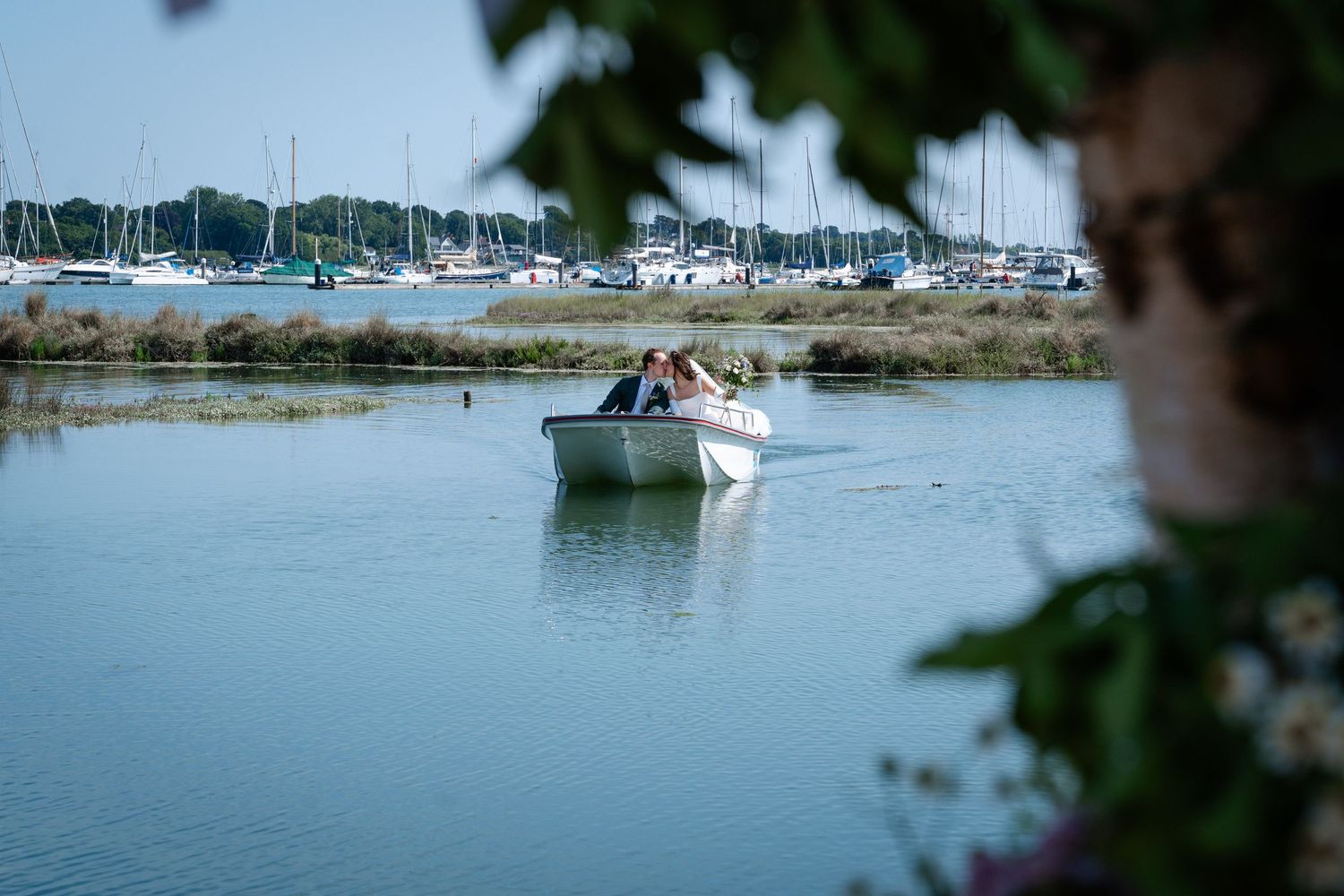 Couple in wedding attire sharing a romantic boat ride on calm water with sailboats and green trees in background.