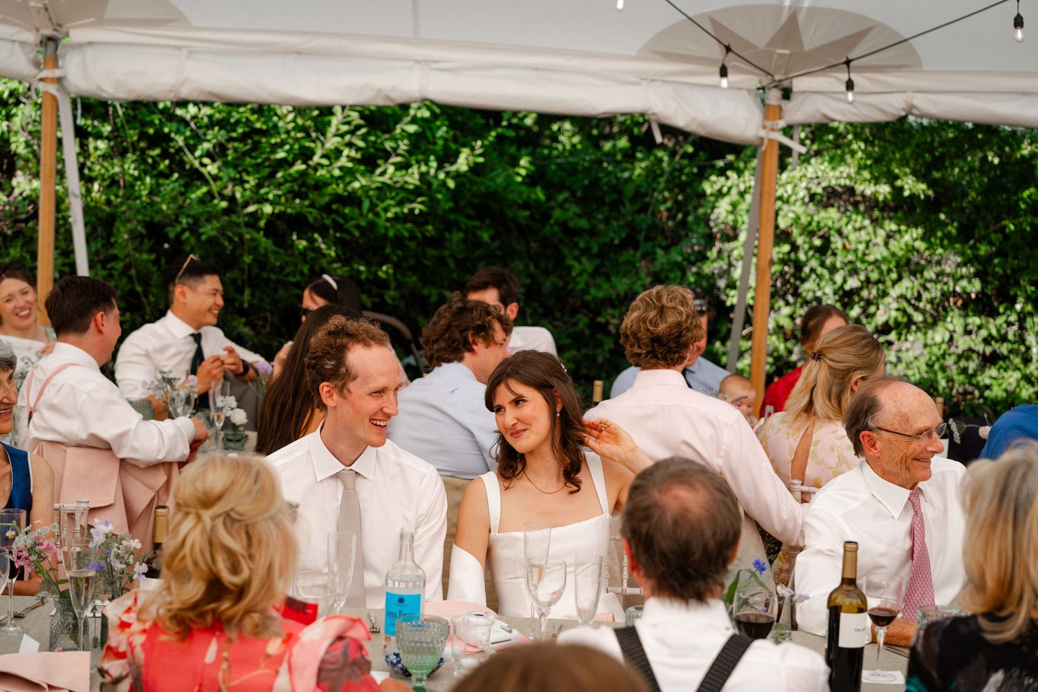 Guests laughing and celebrating at an outdoor wedding reception under a white tent surrounded by lush greenery.