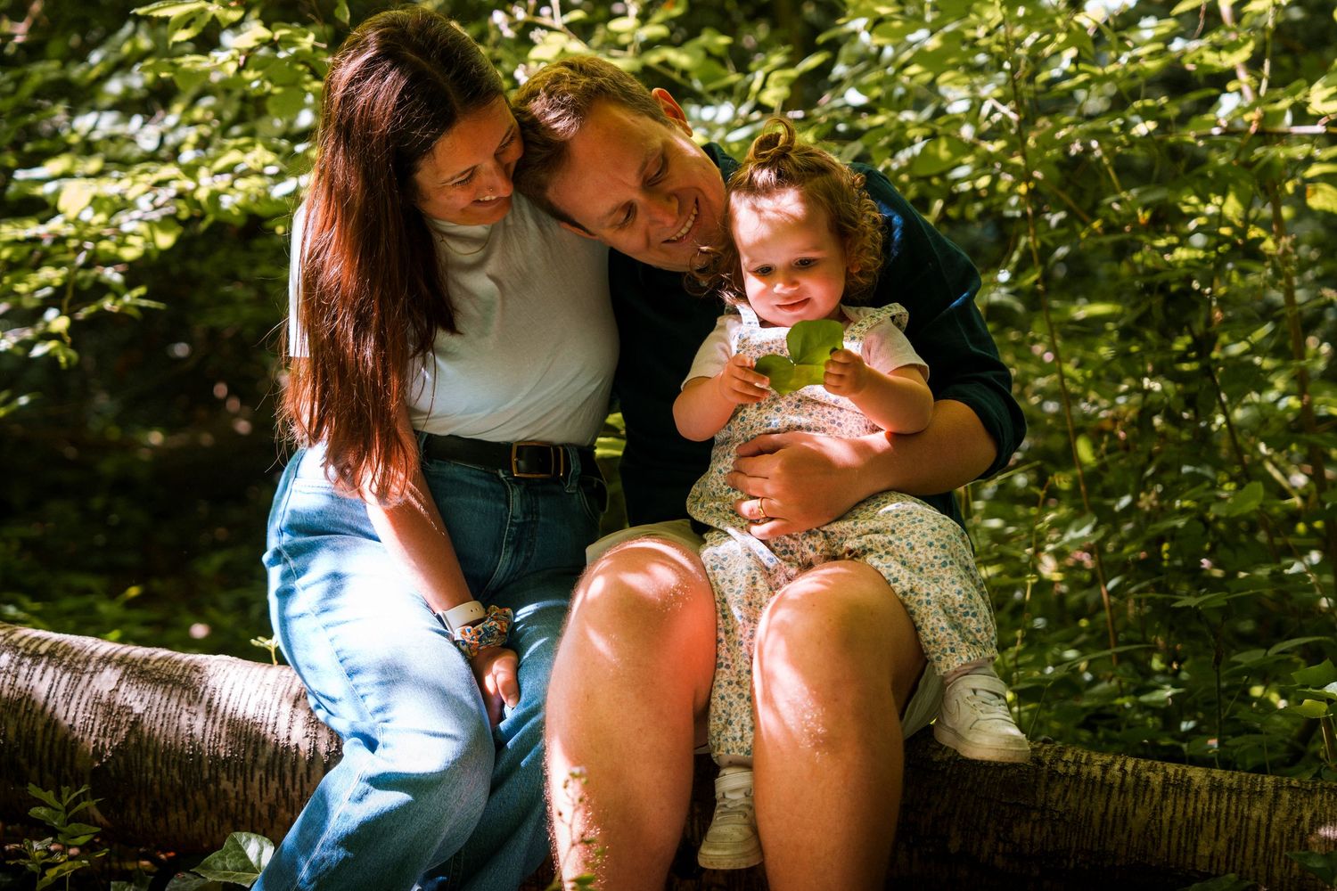 Two parents sitting on a log in a lush green forest, lovingly embracing their young toddler daughter.