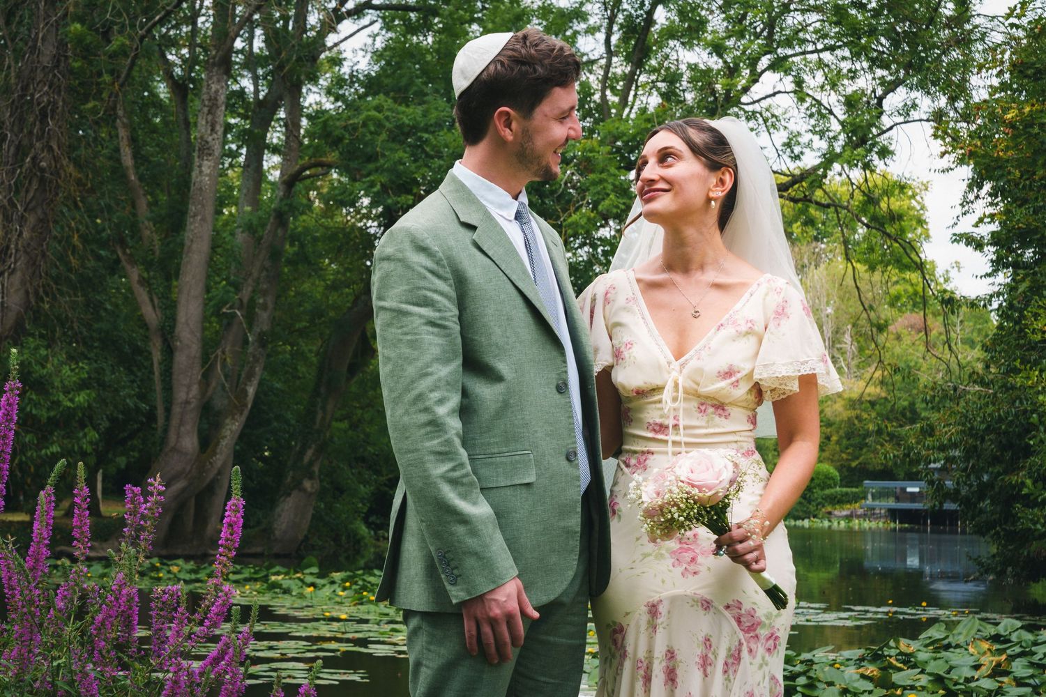 A couple in wedding attire smiles at each other in a lush garden with a pond and lily pads behind them.