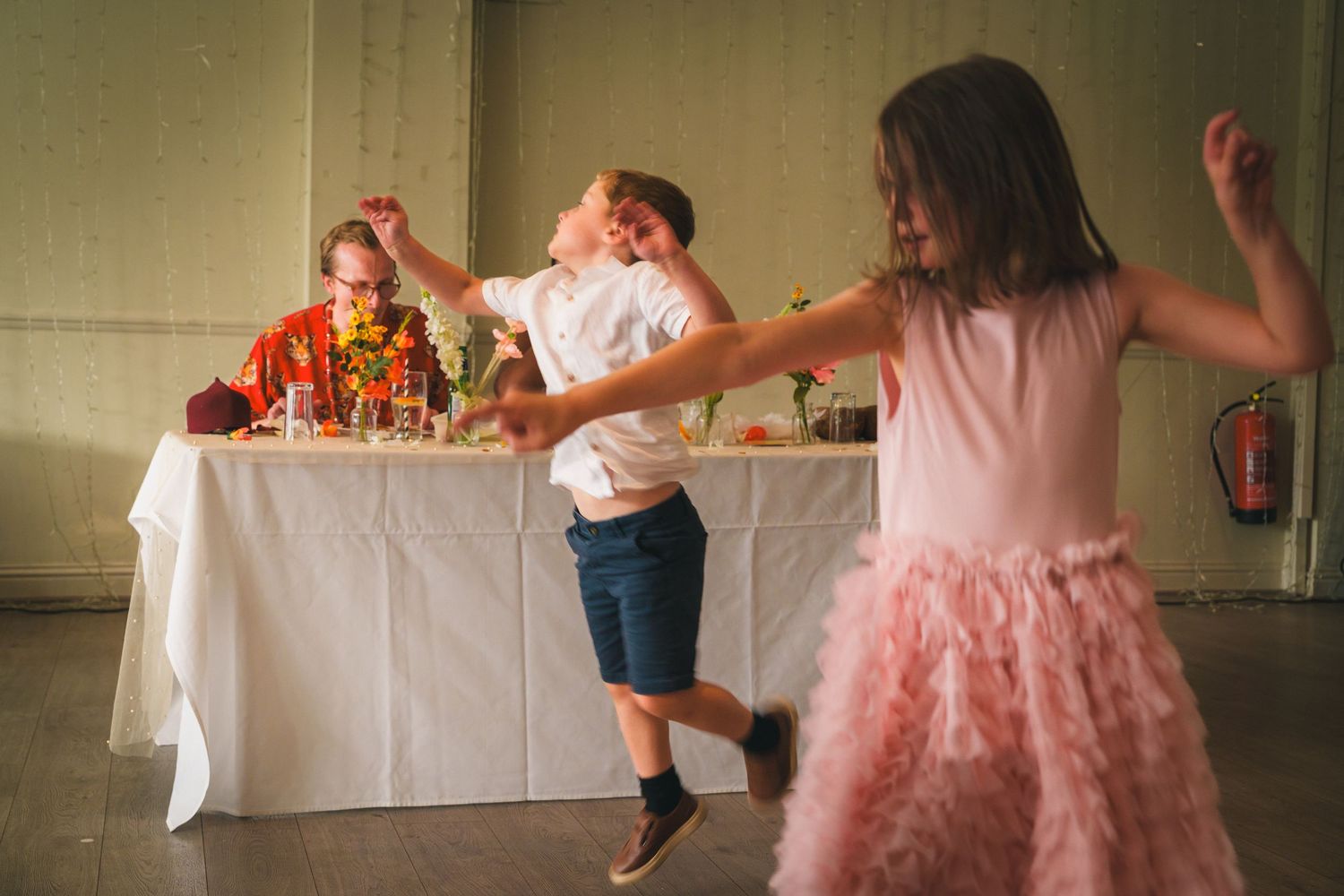 Two children dancing joyfully at a celebration while an adult sits at a decorated table in the background.