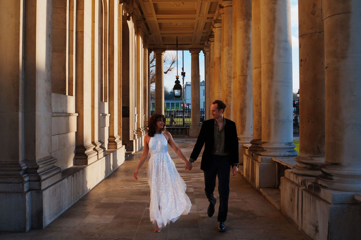 Sunlit colonnade walkway with classical architecture featuring tall stone columns and ornate ceiling details.