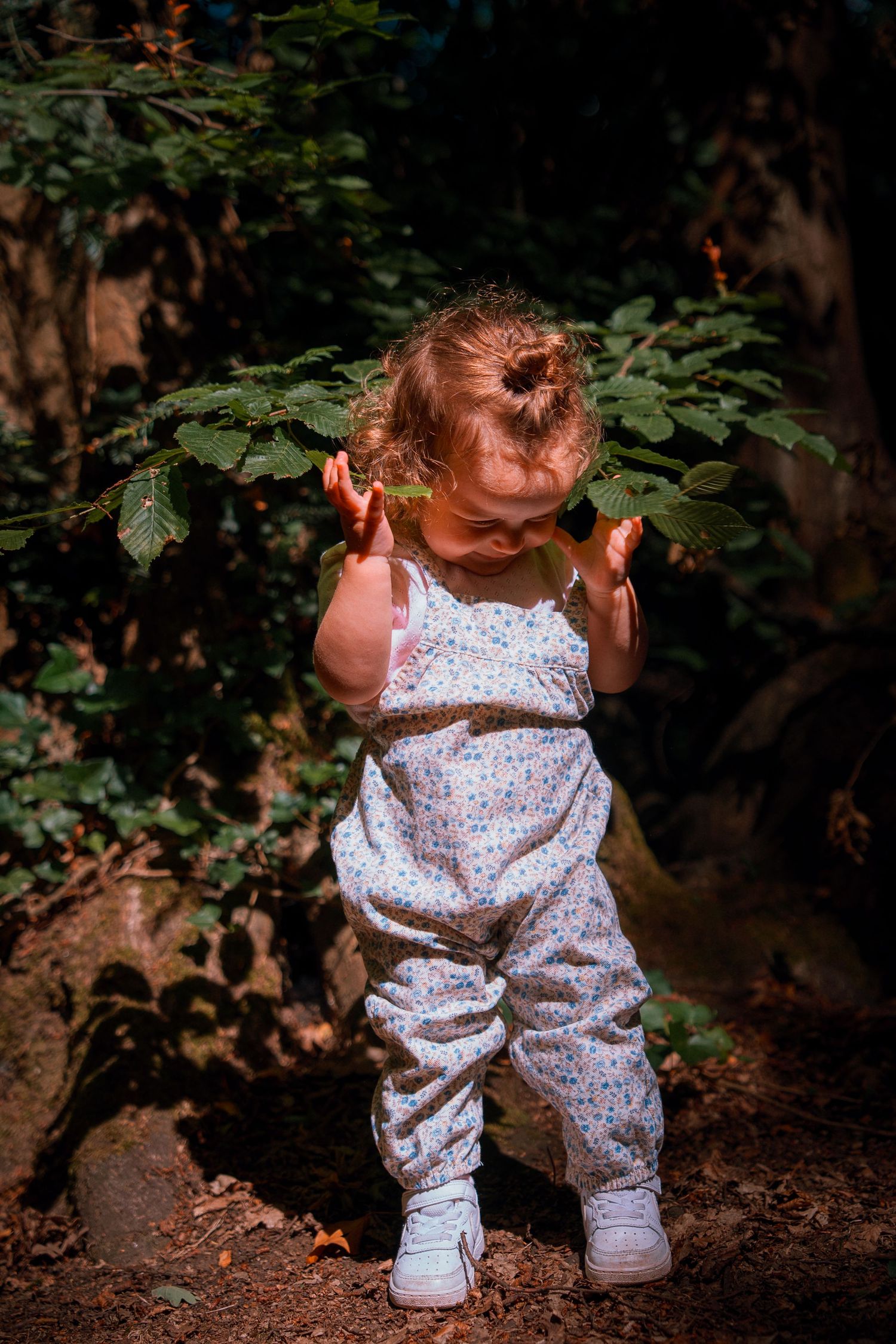 Toddler girl in floral overalls exploring a forest, touching green leaves with both hands.