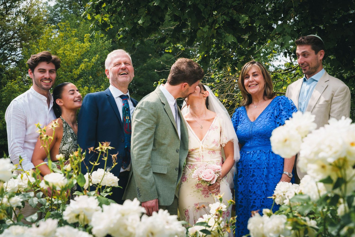 Wedding couple kissing surrounded by family among white flowers in garden ceremony celebration.