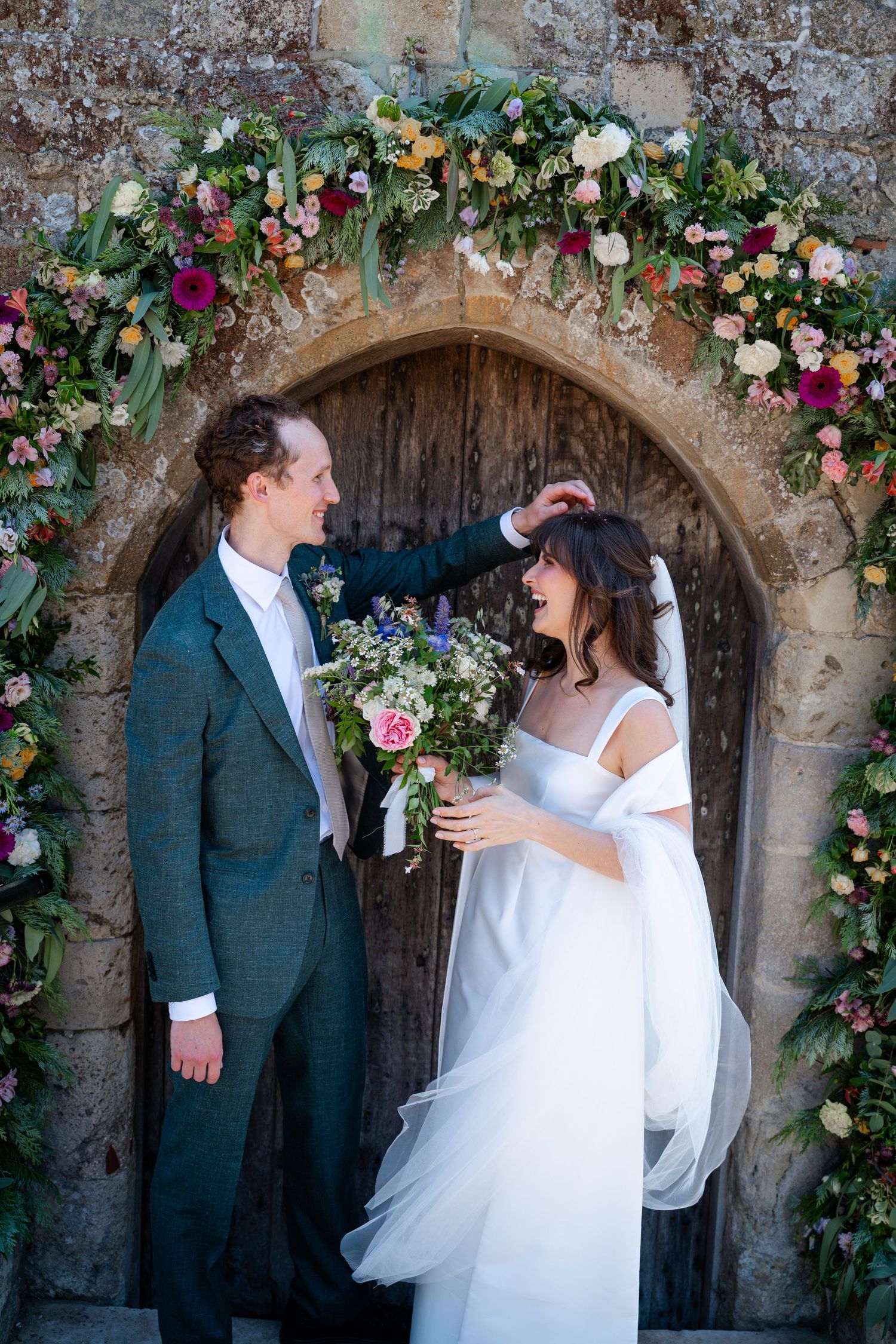 Bride and groom stand beneath a colorful floral arch at a stone church doorway, groom adjusting bride's veil.