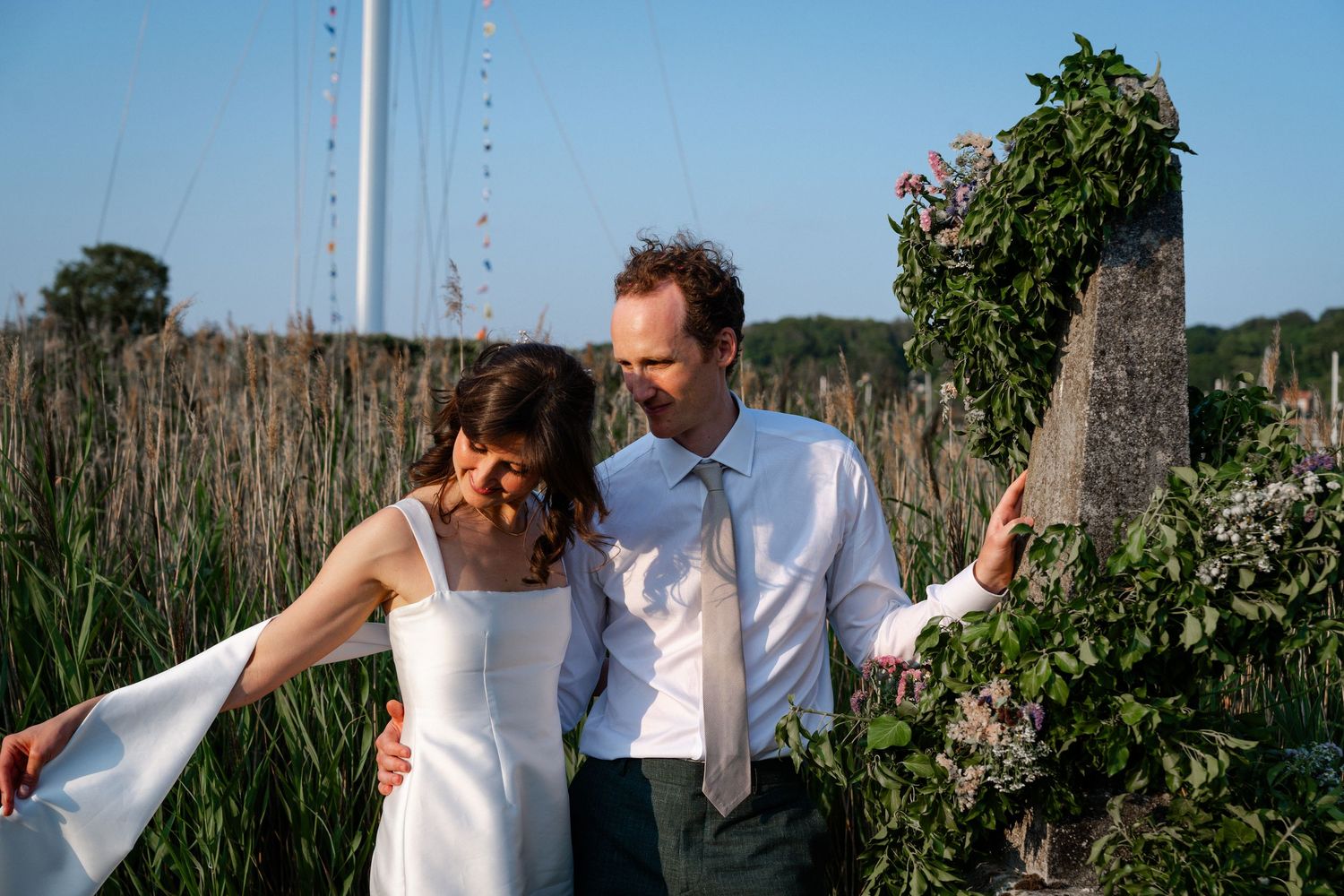 Bride and groom smiling outdoors near a floral maypole, groom in a suit with tie, bride in a white dress.