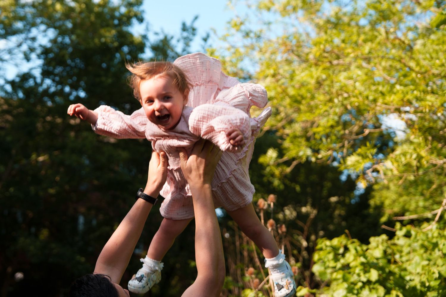 Adult lifting a laughing baby girl in a pink dress high in the air outdoors with green trees in background.