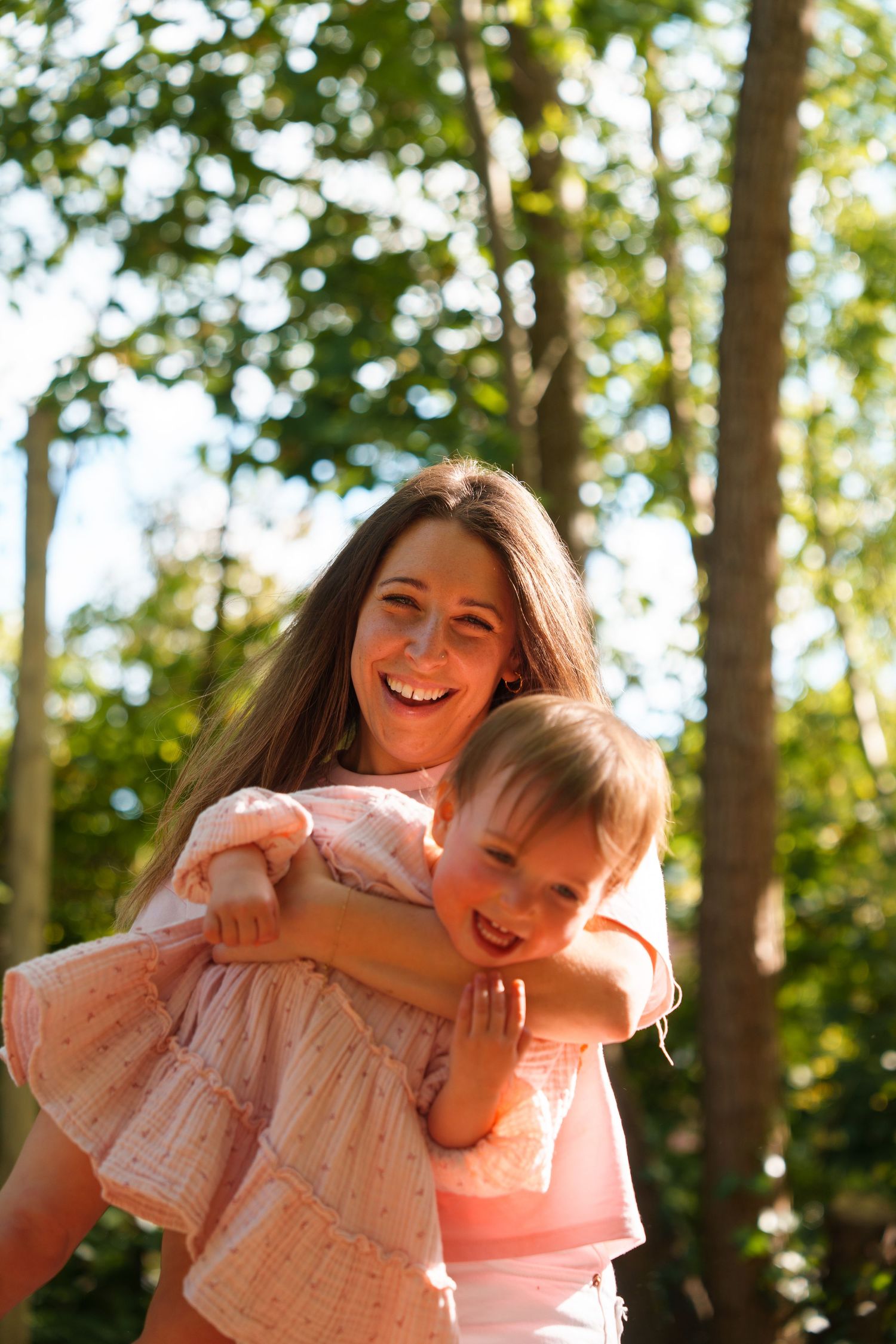 A smiling woman in pink holds a happy toddler girl outdoors in a sunny green park setting.