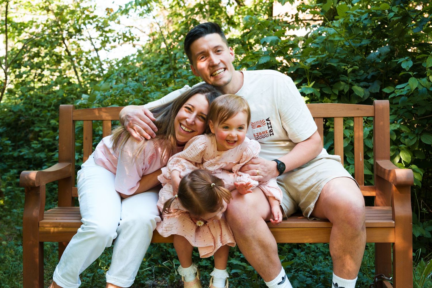 Happy family of four sitting on a wooden bench outdoors, smiling together surrounded by lush green trees.