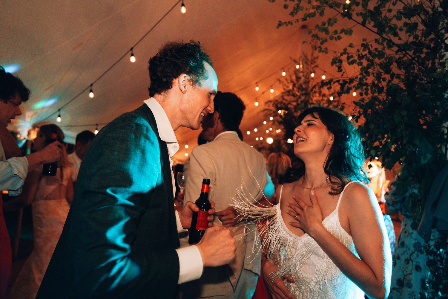 Couple sharing a joyful dance at a wedding reception under warm string lights surrounded by guests.