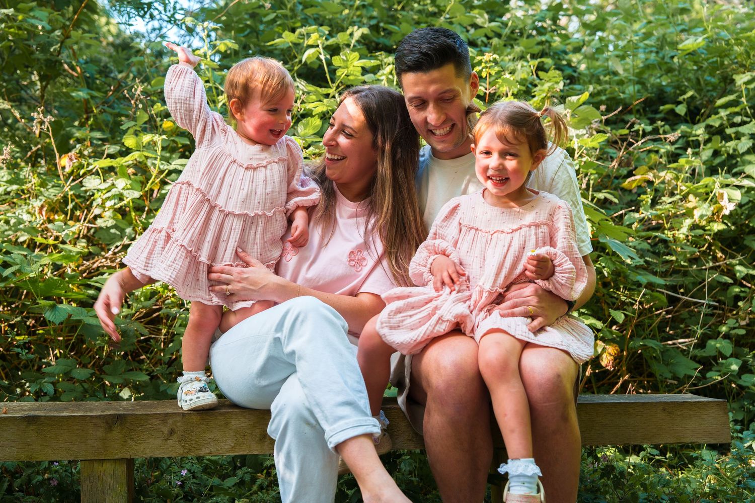 Happy family of four sitting on a bench outdoors, parents with two young girls in matching pink dresses, surrounded by greenery.