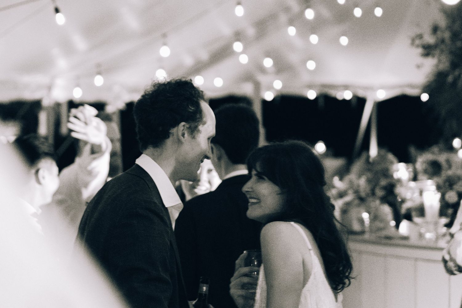 Black and white photo of a bride and groom sharing a romantic moment under string lights at their wedding reception.
