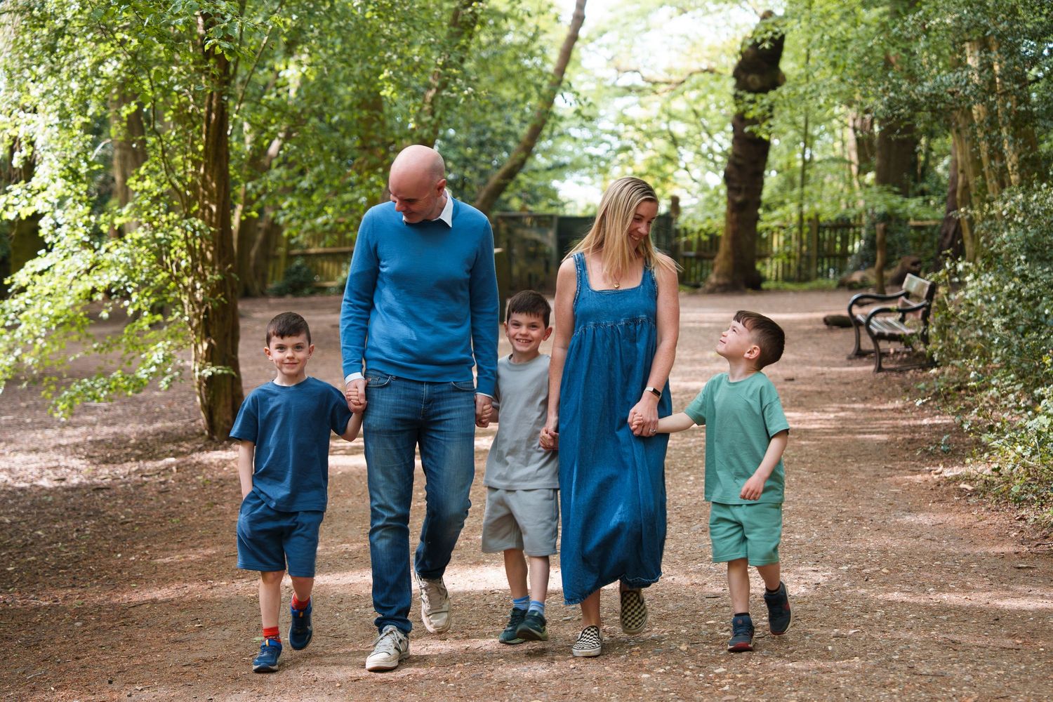 Family of five walking together on a woodland path, parents holding hands with three young boys among lush green trees.
