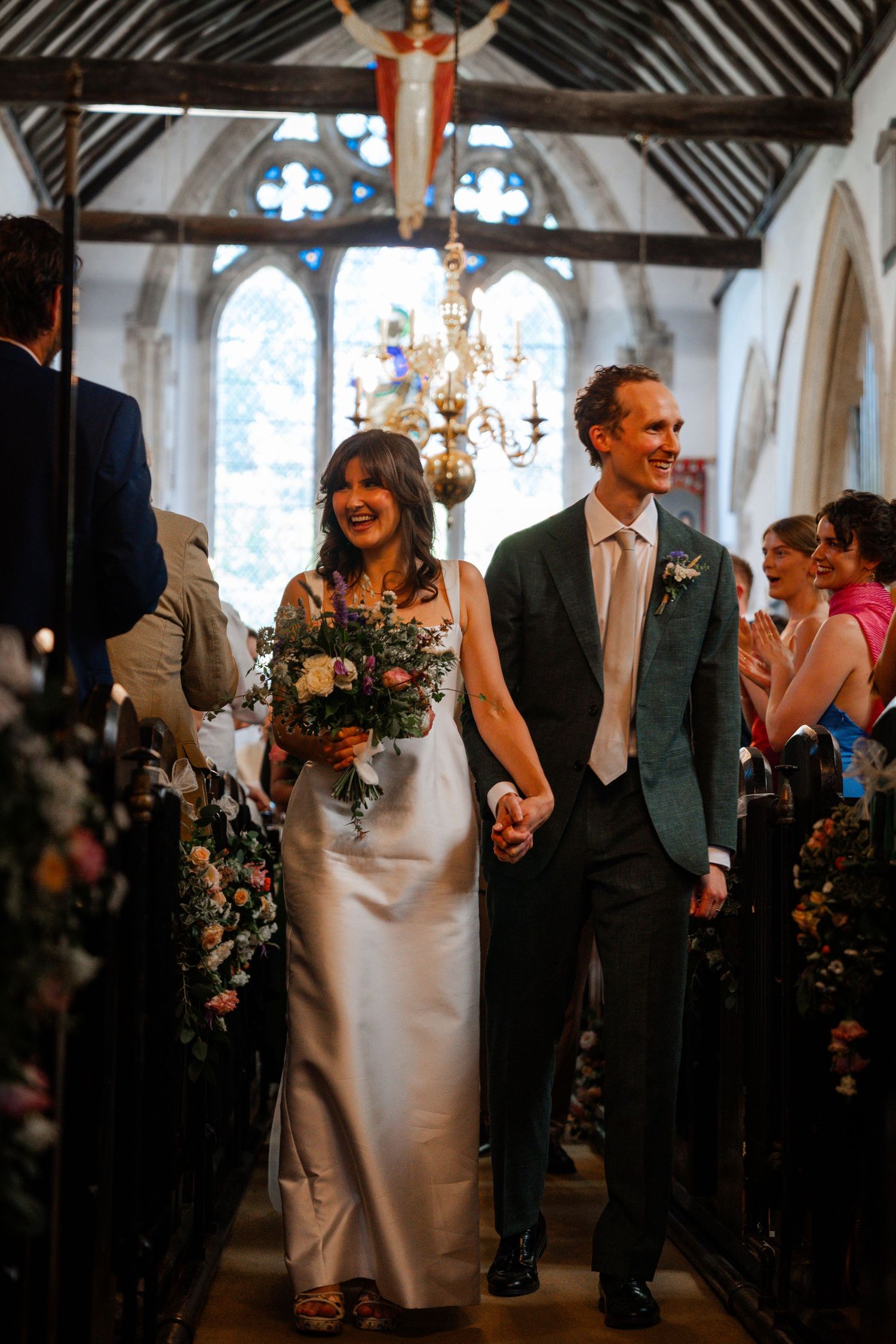 Bride and groom walk hand-in-hand down church aisle, smiling joyfully, bride holding floral bouquet in elegant satin gown.