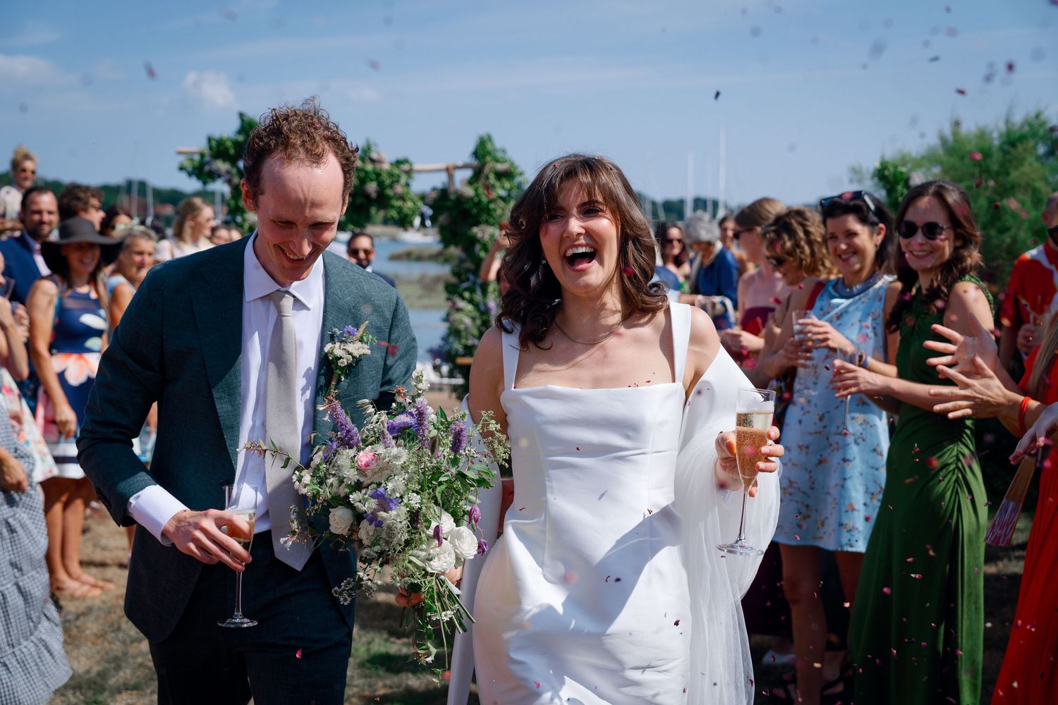 Joyful bride and groom walk through confetti shower at outdoor waterfront wedding ceremony on sunny day.