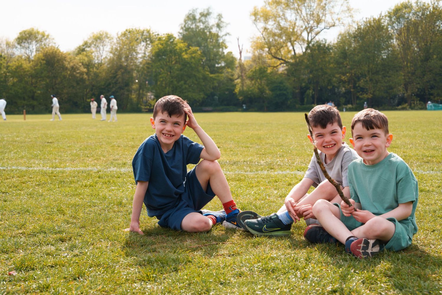 Three young boys sitting on green grass at a cricket field, smiling at the camera on a sunny day.