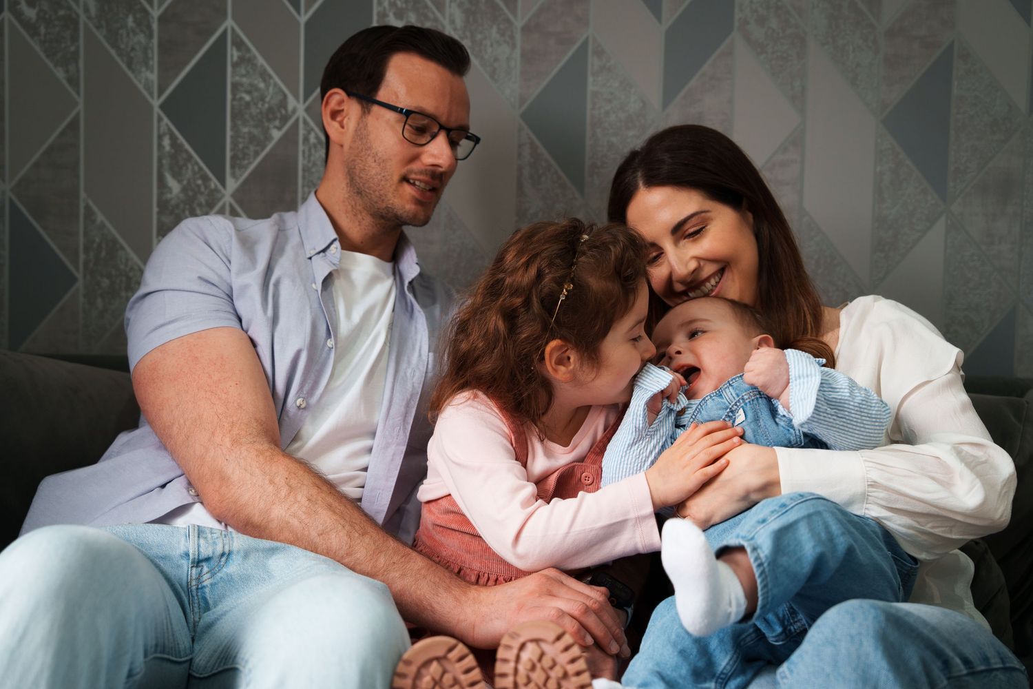 Happy family portrait with parents and two children sitting together on a couch, wearing casual denim and white clothing.