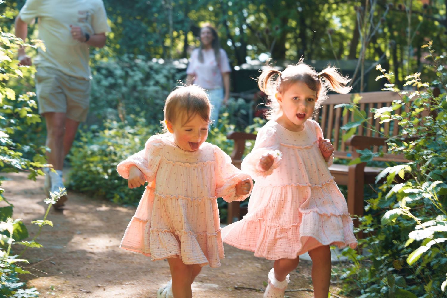 Two young girls in matching pink dresses running happily along a garden path surrounded by lush green trees.