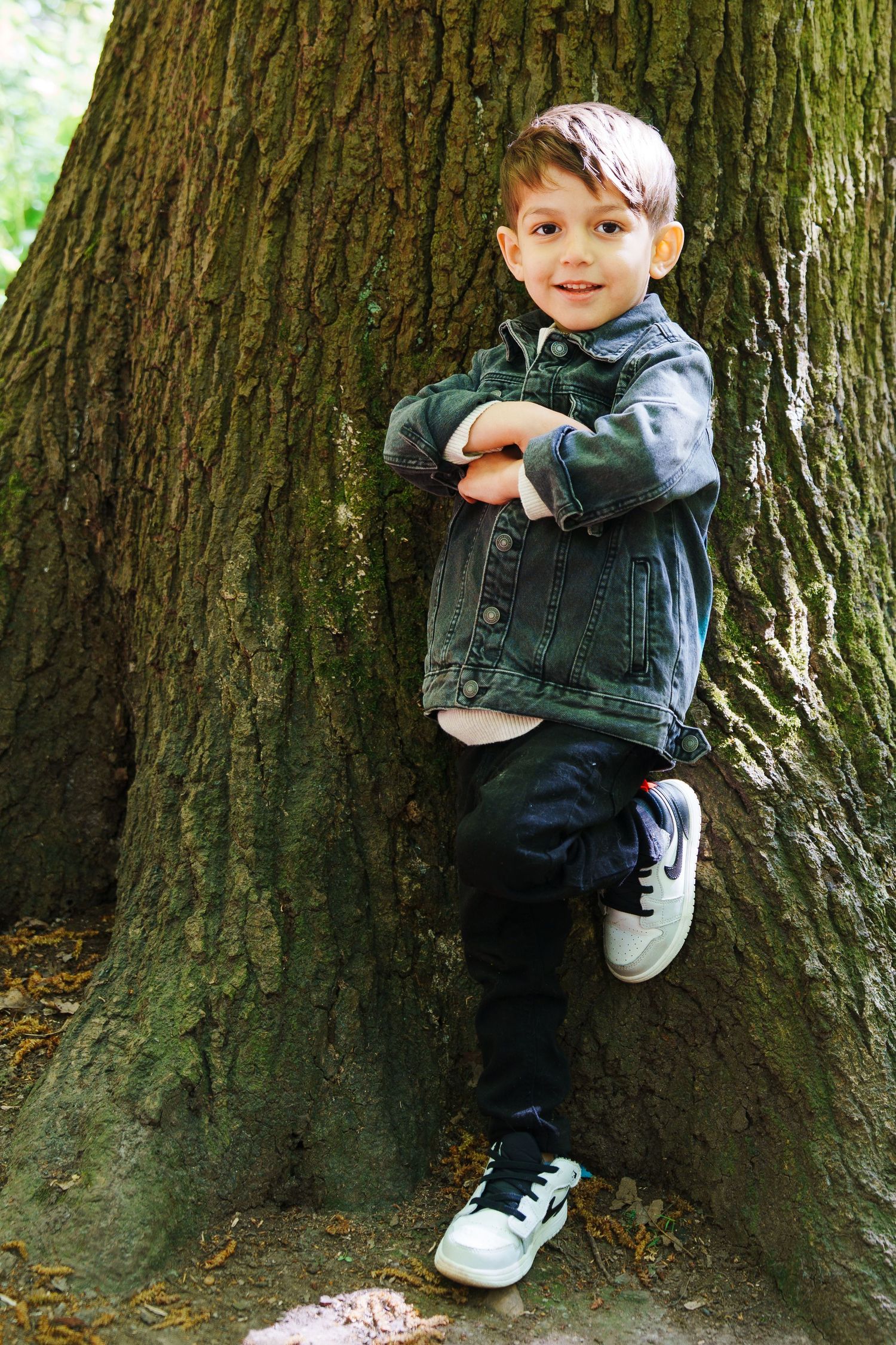 Young boy in denim jacket leaning against large tree trunk with arms crossed, smiling outdoors.