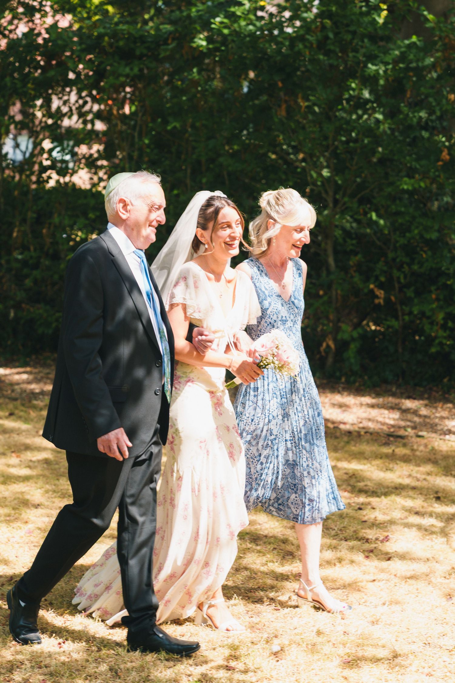 Bride walks with parents on sunny wedding day, laughing joyfully in elegant white gown and veil outdoors.