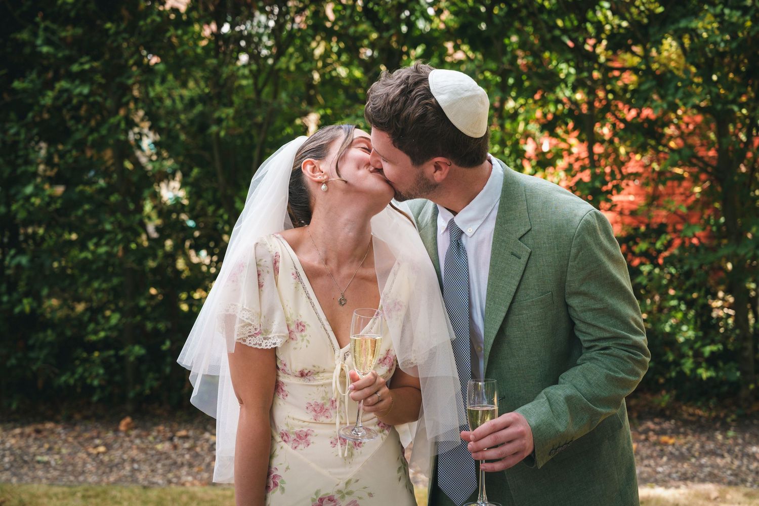 Bride and groom sharing a kiss while holding champagne glasses after Jewish wedding ceremony outdoors.