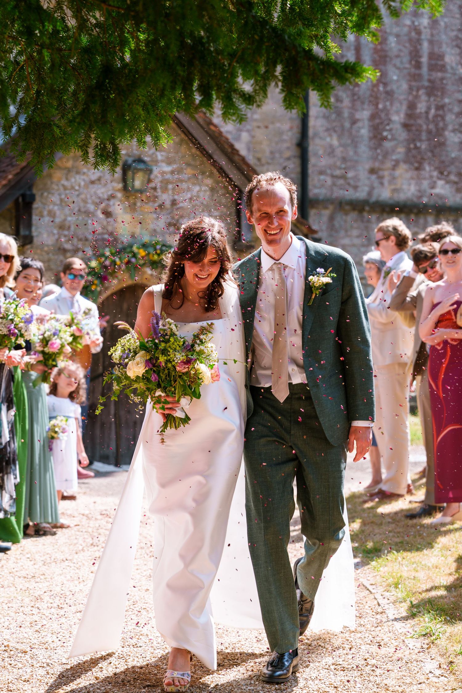 Bride and groom walk through confetti shower outside stone church, bride holding wildflower bouquet in white gown.