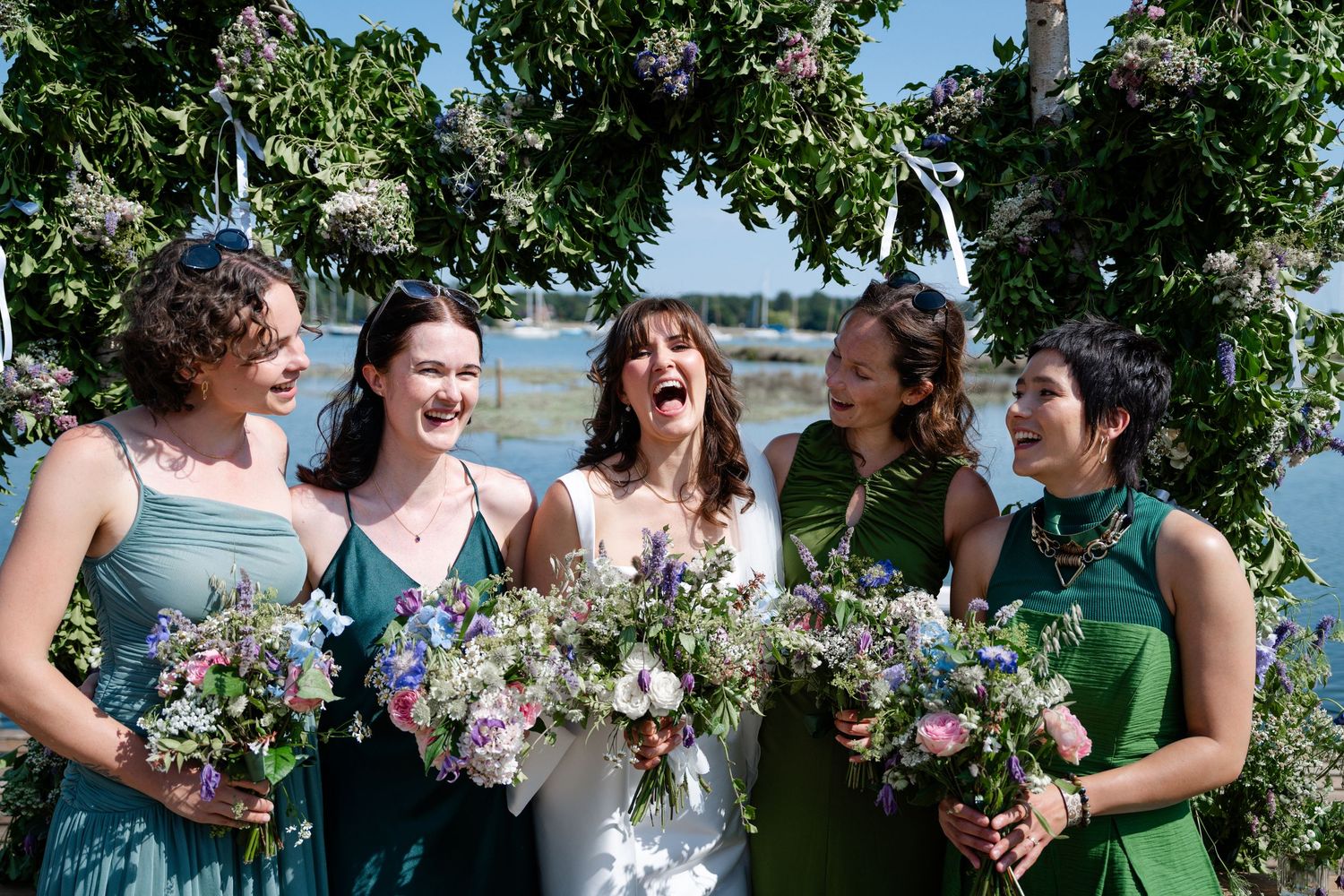 Bride laughing with four bridesmaids in green dresses holding wildflower bouquets outdoors near water.