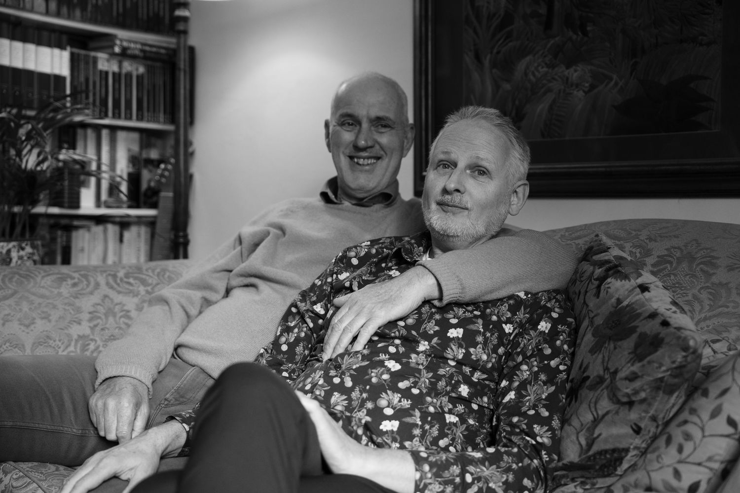Black and white photo of an elderly couple smiling and embracing on a couch in a cozy living room.
