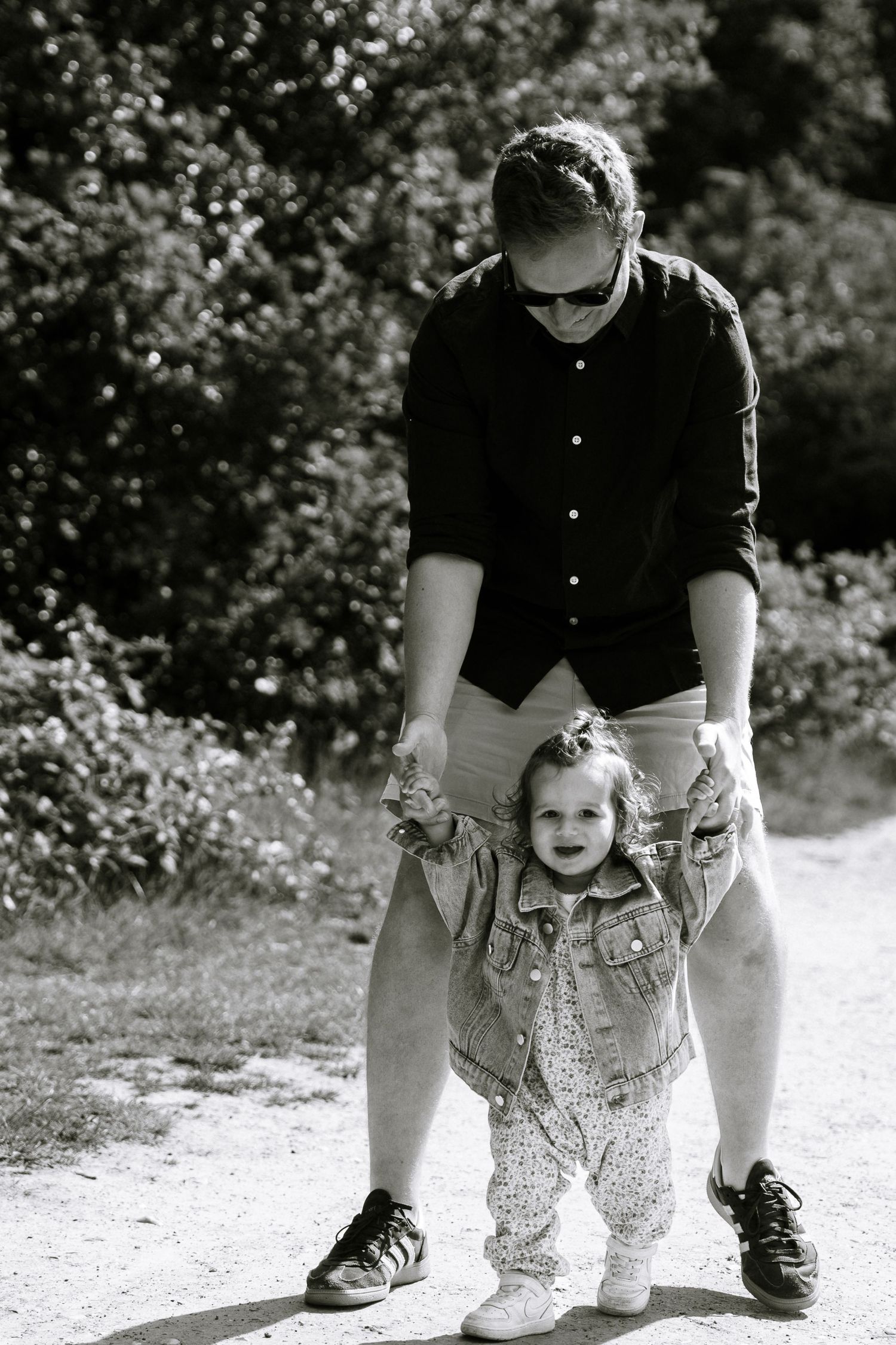 Black and white photo of a parent helping a toddler walk on a sunny outdoor path surrounded by trees.
