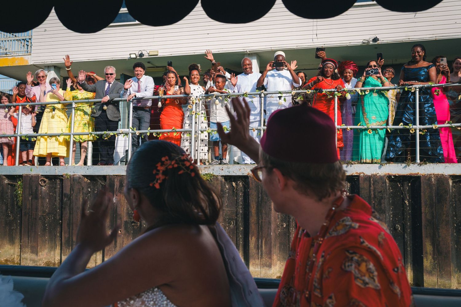 Colorful wedding guests wave from a decorated balcony as two people in festive attire wave back below.