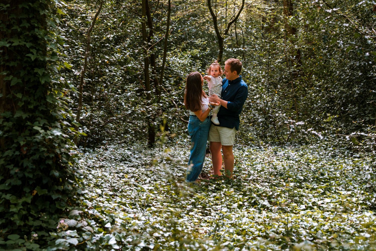 Family of three standing among white wildflowers in a lush green forest, parents holding young child.