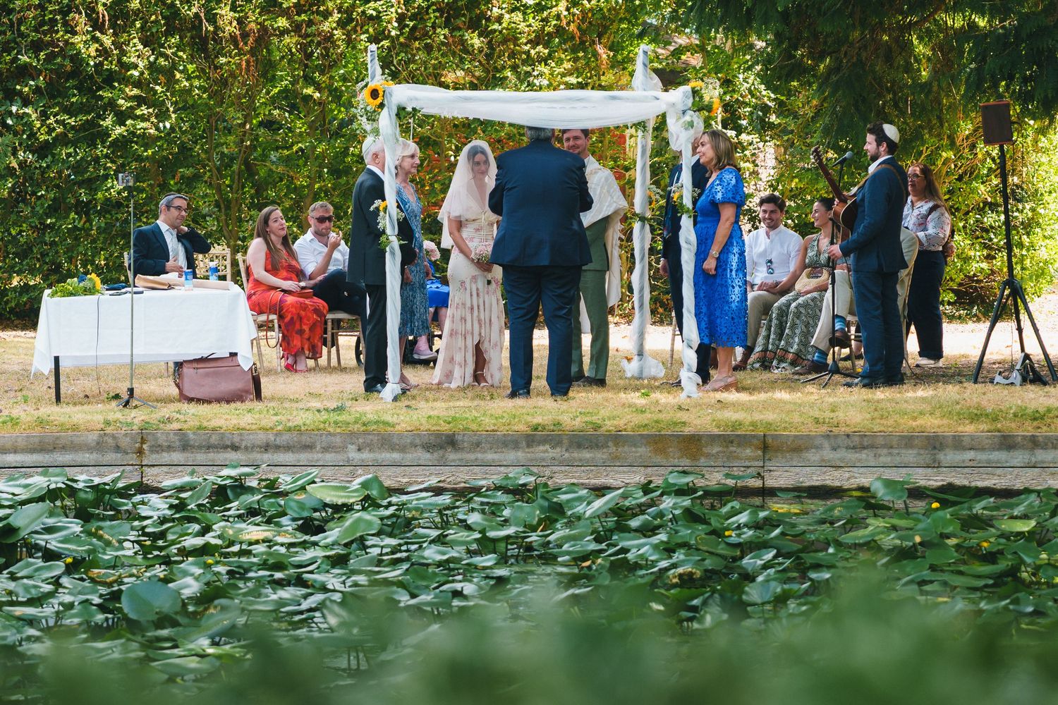 Outdoor wedding ceremony under floral chuppah beside a lily pond, surrounded by seated guests and lush green trees.