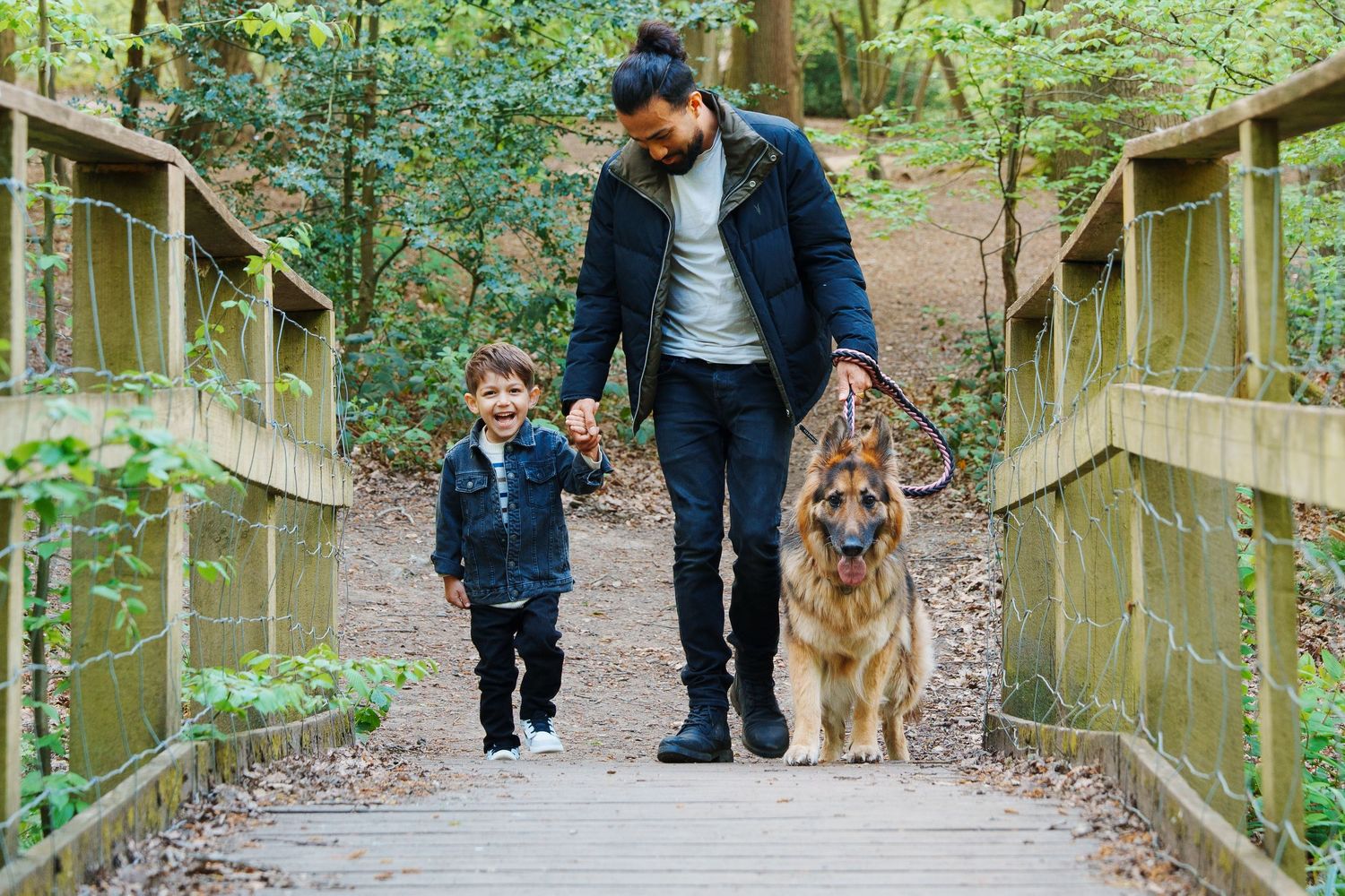 Man and child walk a German Shepherd dog across a wooden bridge in a lush green forest.