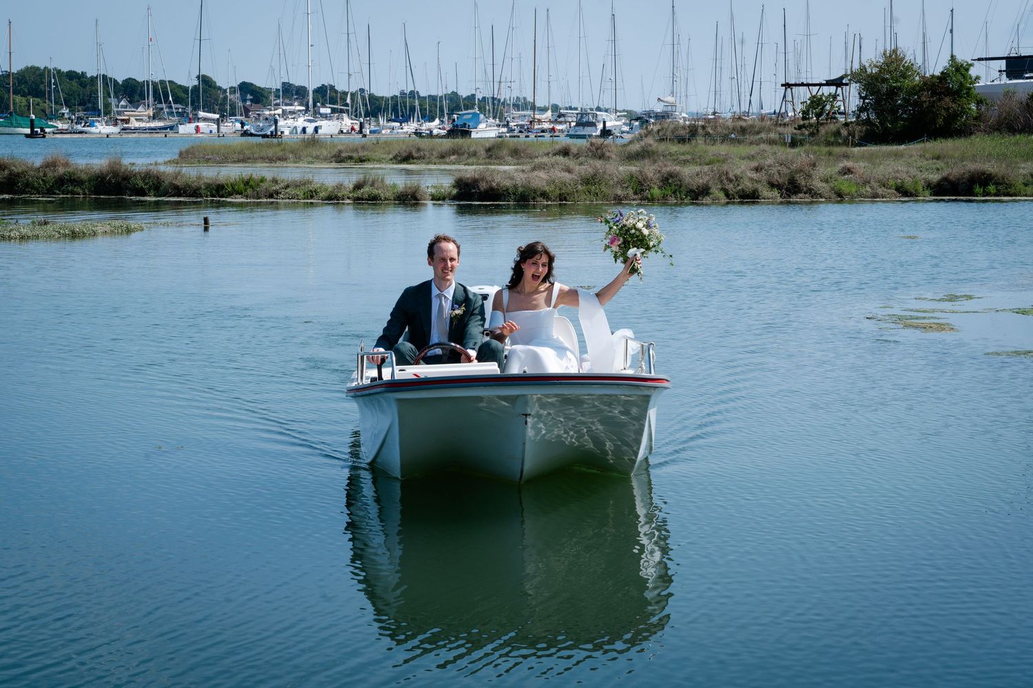 Bride and groom riding a small white motorboat on calm water with a marina in the background on their wedding day.