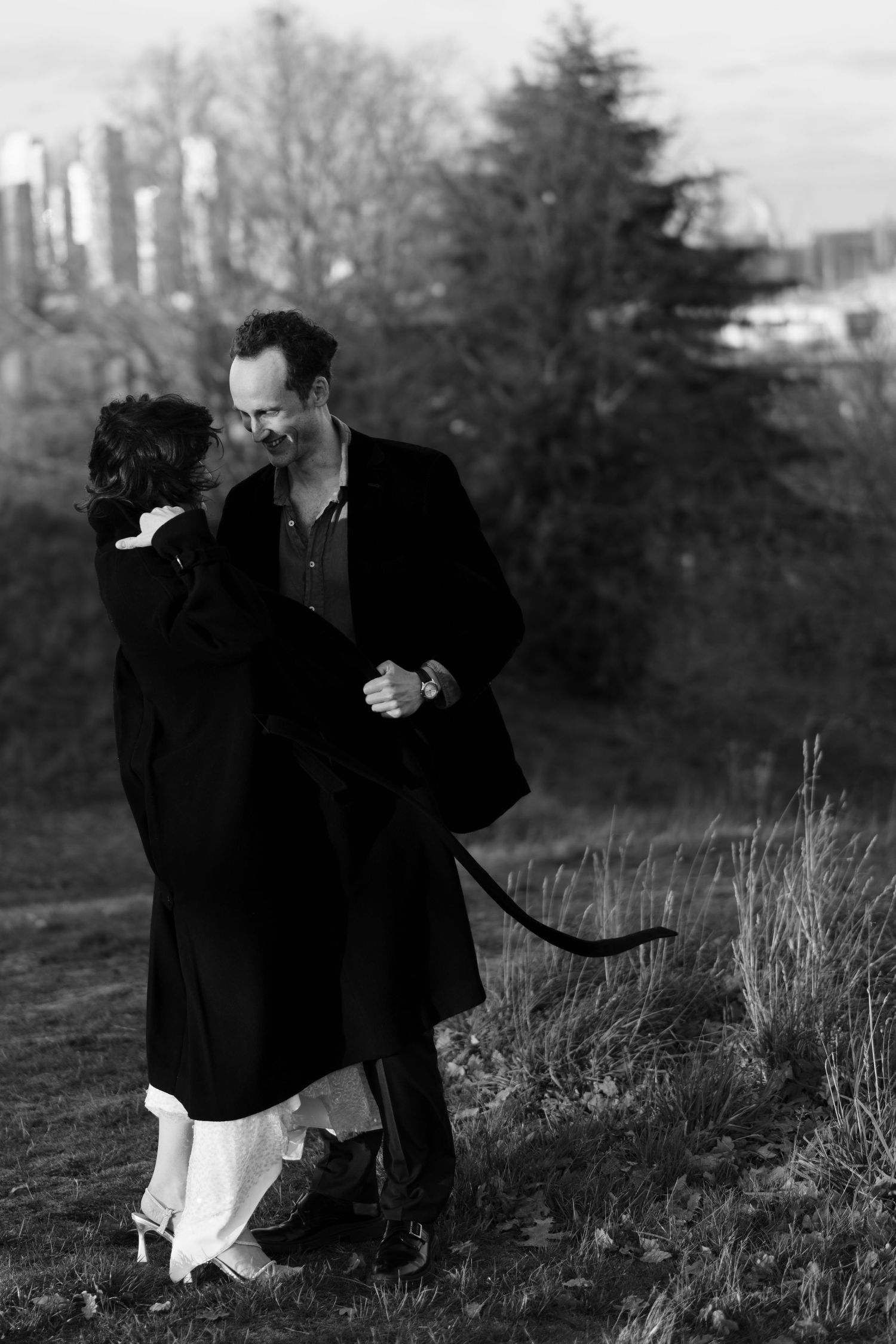 A black and white image of a couple sharing an intimate moment outdoors with urban buildings visible in the distance.