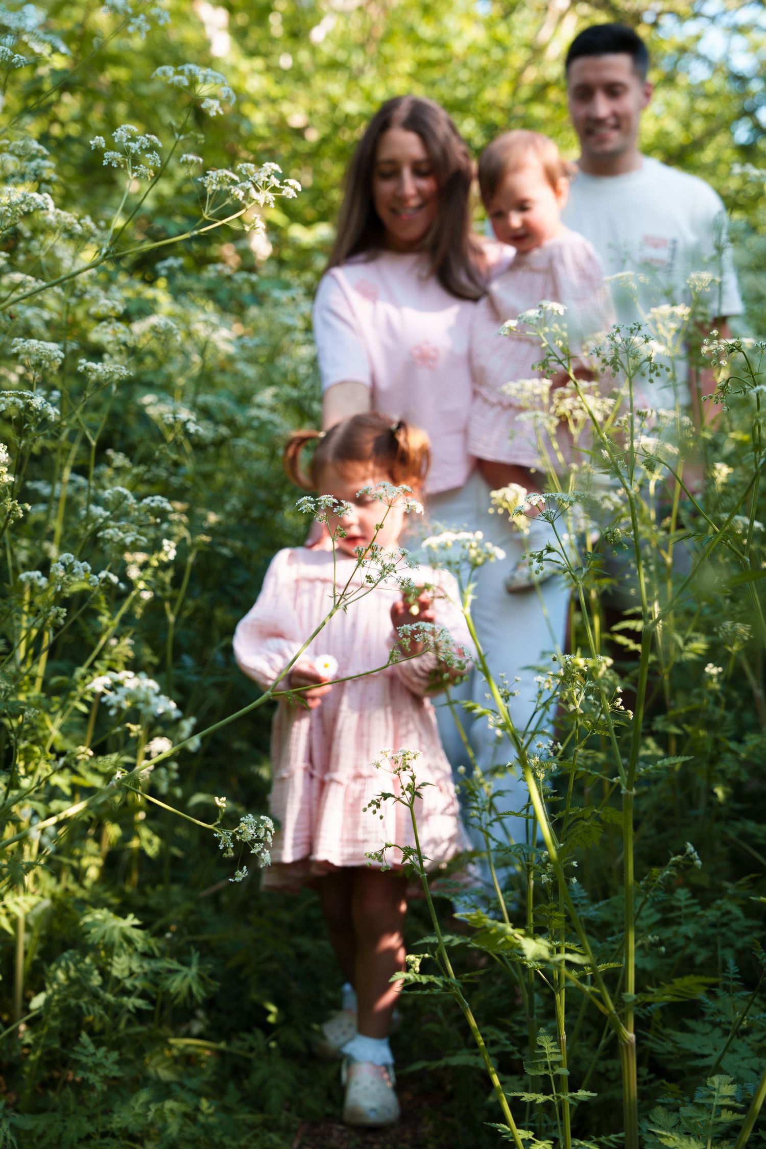 Family with two young girls in pink dresses walking through lush green wildflower meadow on sunny day.
