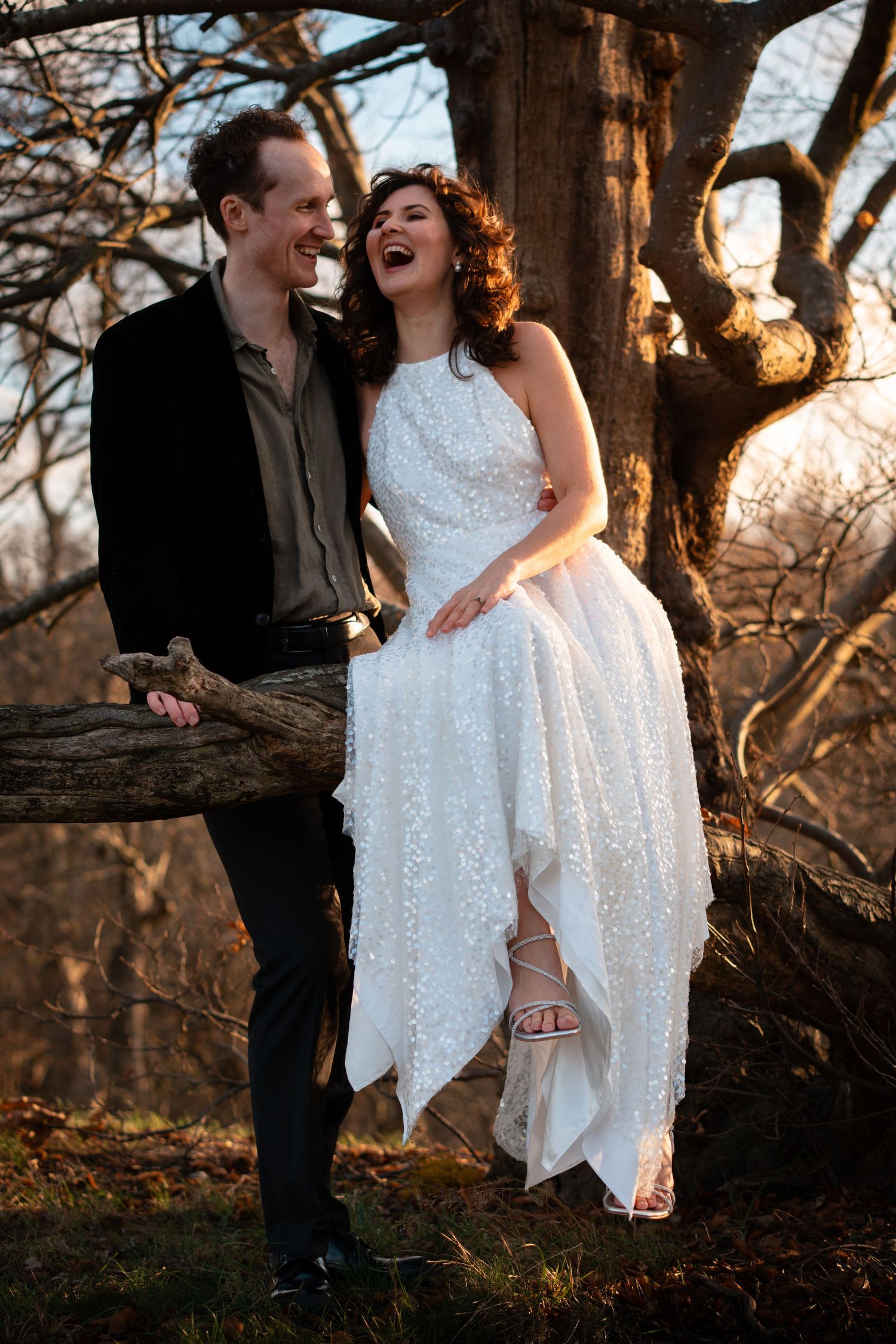 A joyful couple laughs together under a tree at sunset, with the bride wearing a sparkly white dress.