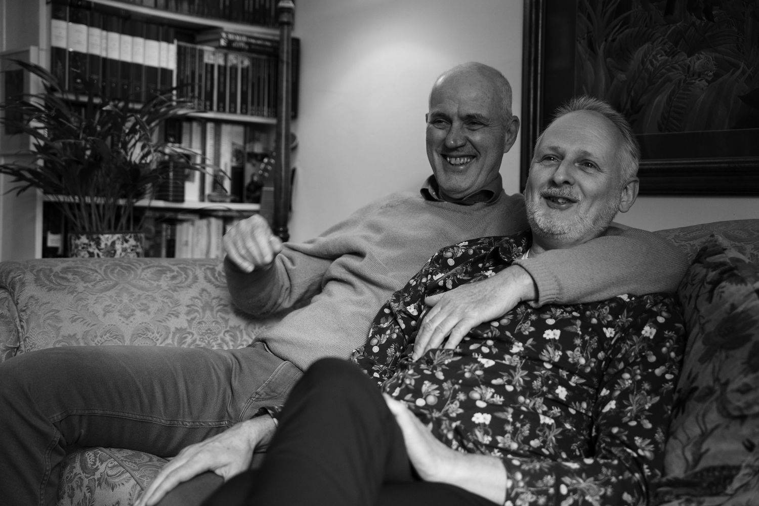 Black and white photo of two smiling men relaxing together on a couch in a cozy living room.