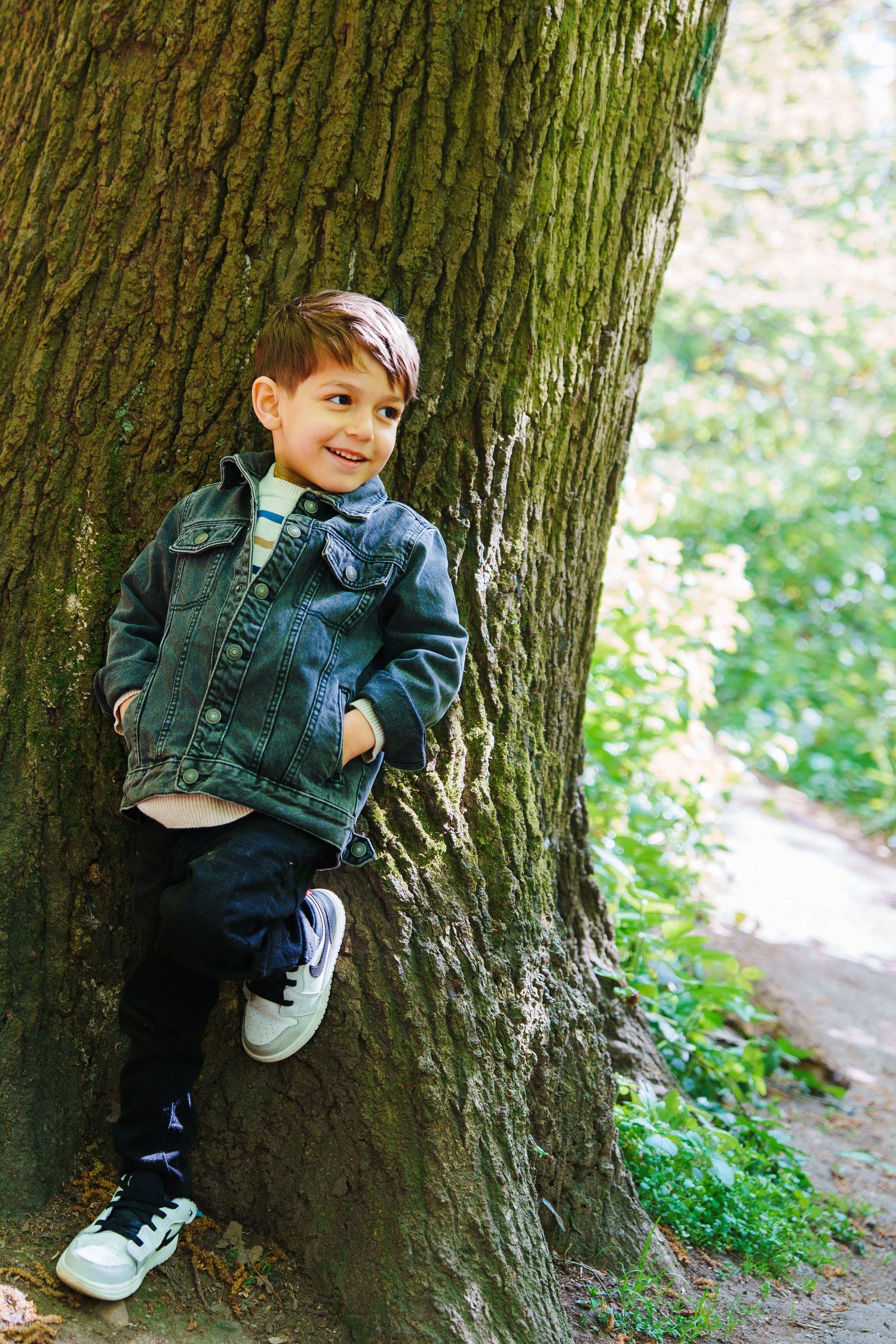 A smiling young boy in a denim jacket leans against a large tree trunk in a lush green park.